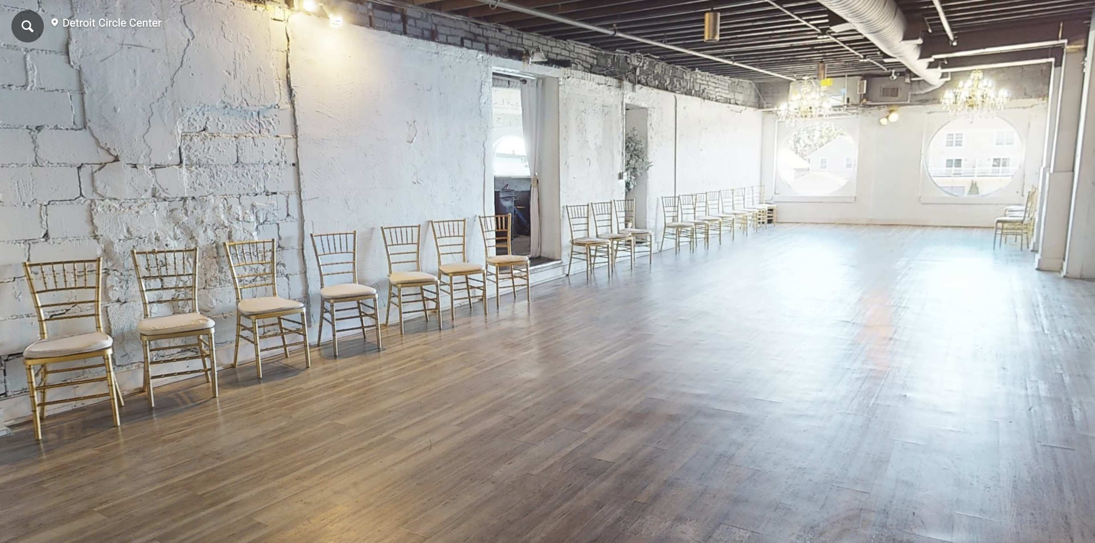 A spacious, empty room with light-colored walls and a wooden floor, featuring rows of gold chiavari chairs along the sides.