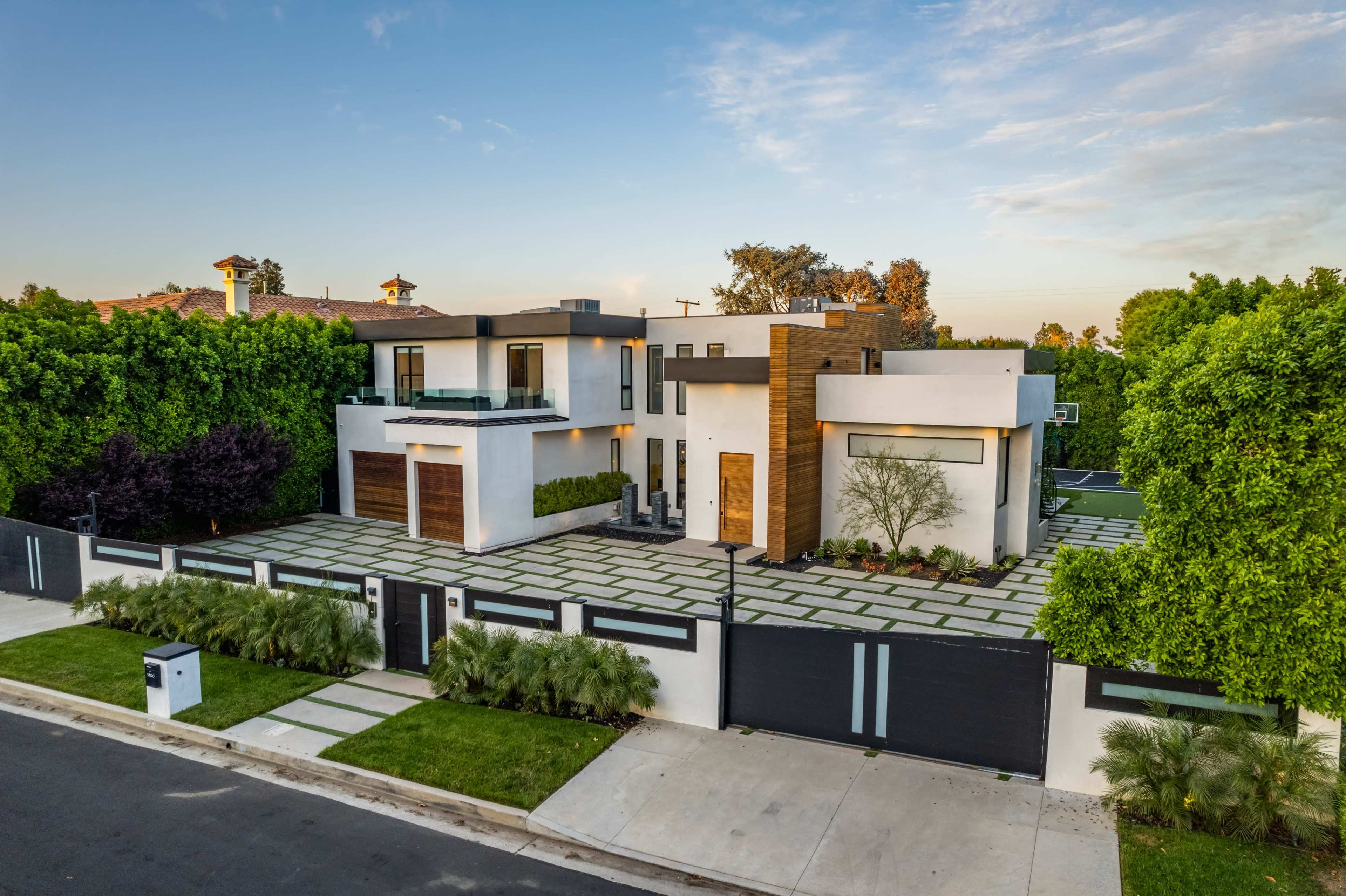 A modern two-story house with a mix of white and wood paneling is surrounded by lush greenery and features a structured driveway.