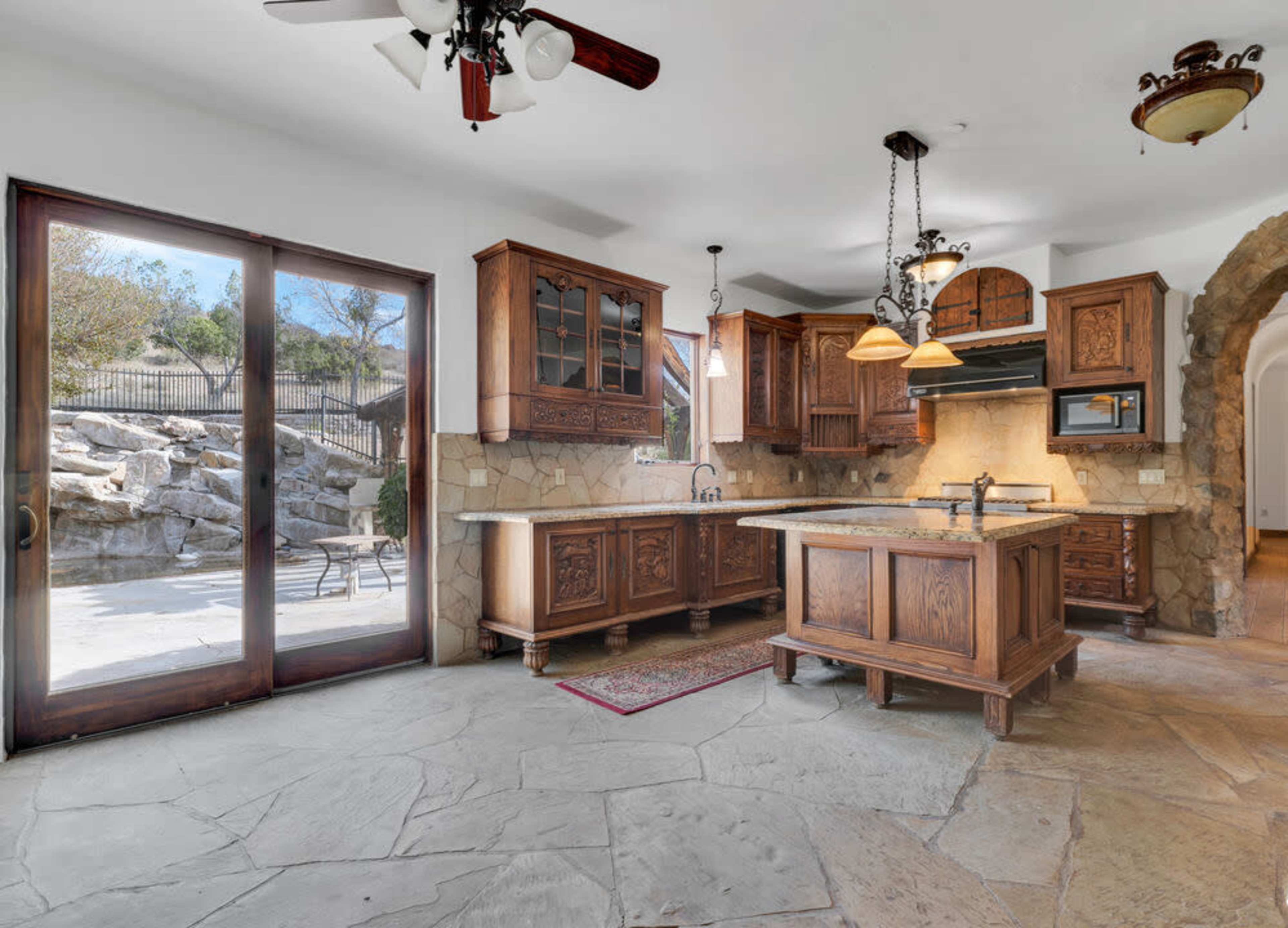 The kitchen features wooden cabinets, a central island with a sink, and stone flooring, with a sliding glass door opening to an outdoor patio area.