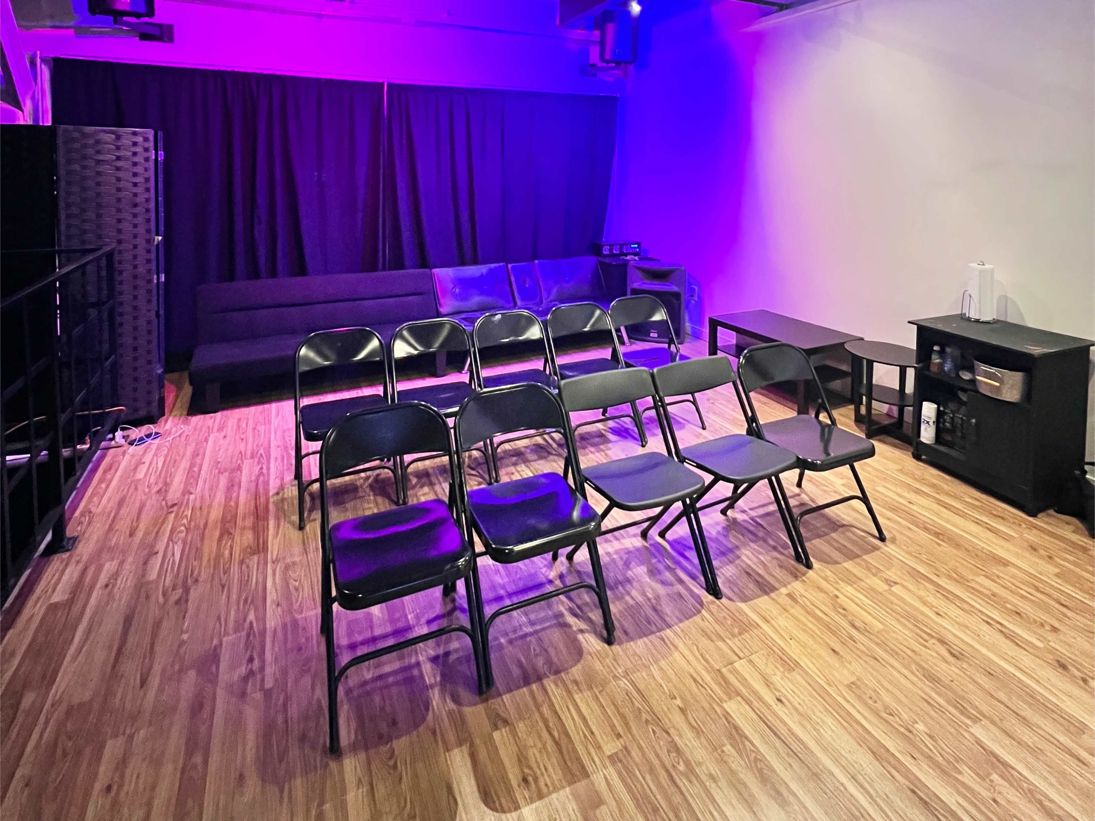 The room features a set of black folding chairs arranged in rows, a dark sofa along one wall, and tables on the other side, with purple lighting illuminating the space.
