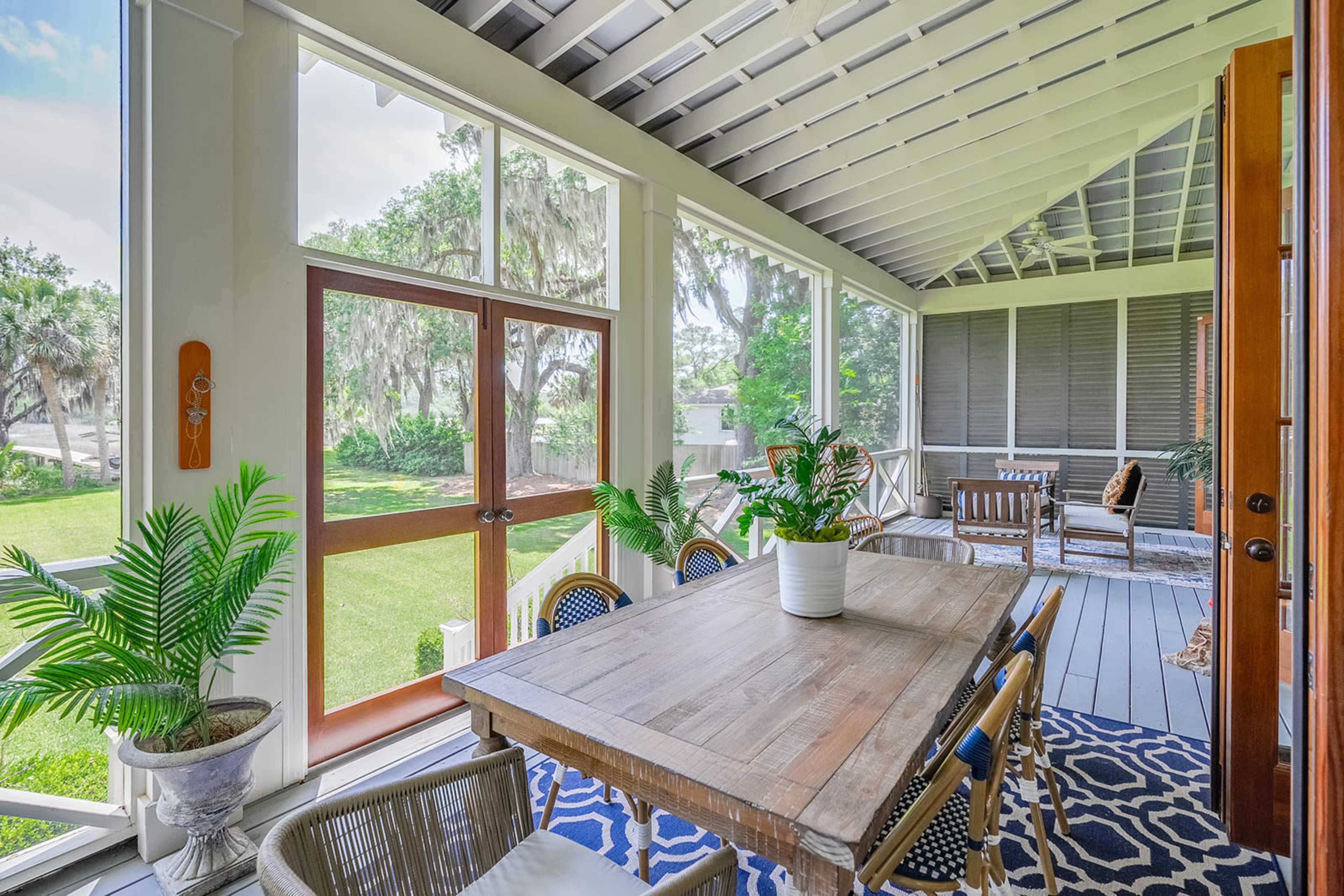 A screened porch features a wooden dining table surrounded by chairs, with potted plants and a view of a green lawn through large windows.