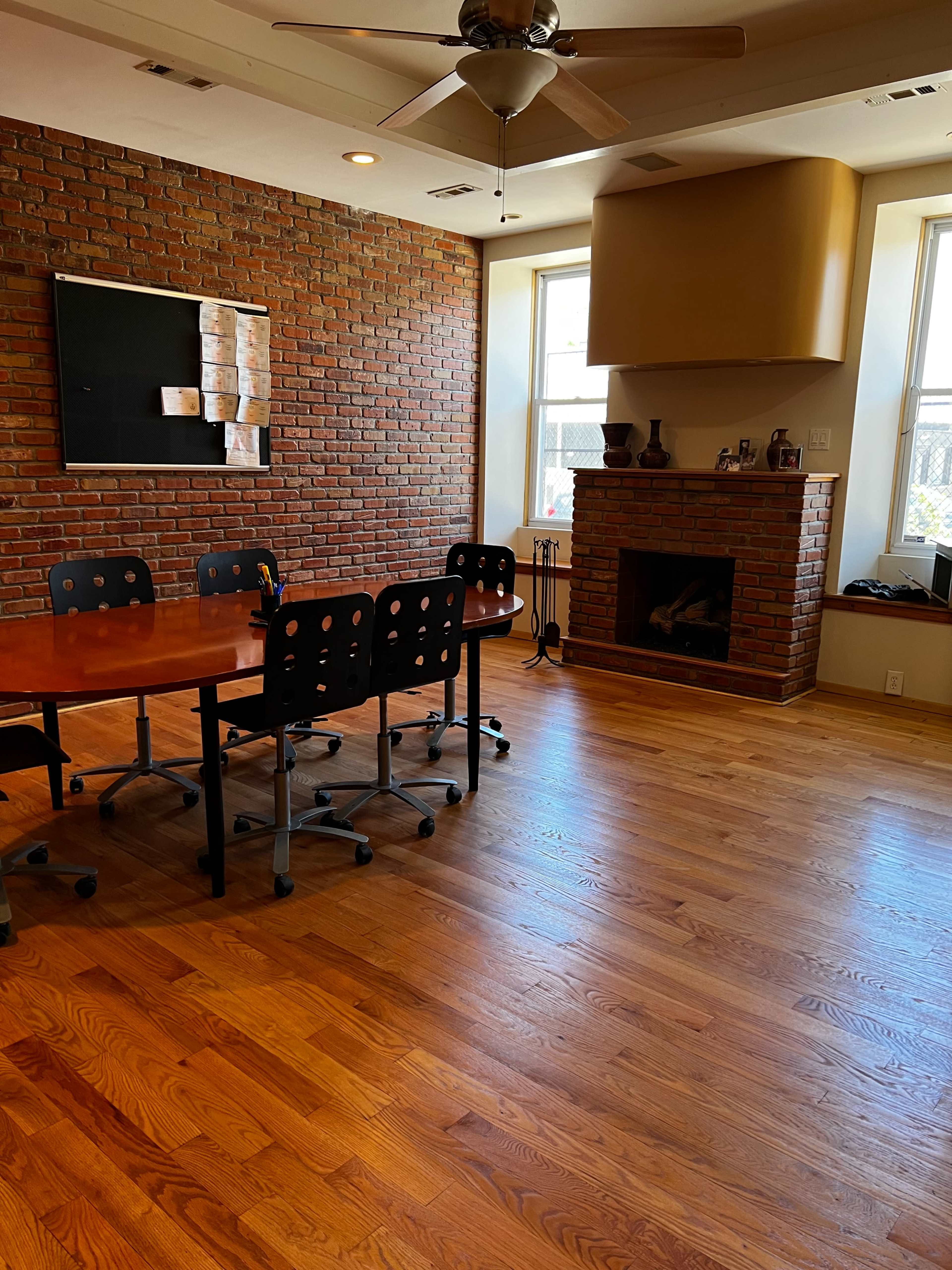 A meeting room with a round wooden table and black rolling chairs, complemented by a brick wall and a fireplace.