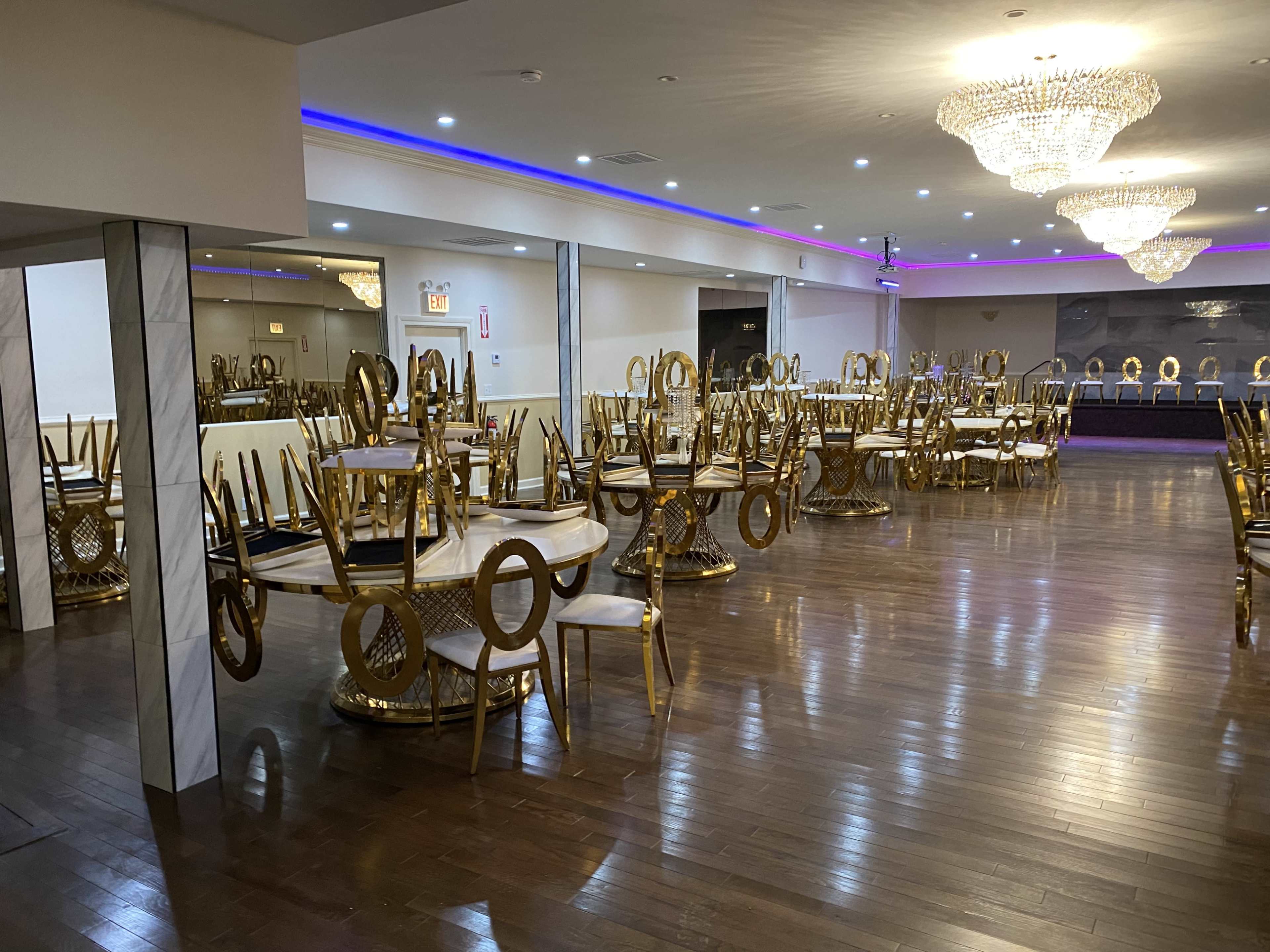 An empty banquet hall with several gold-accented dining tables and chairs stacked upside down, along with decorative chandeliers overhead.