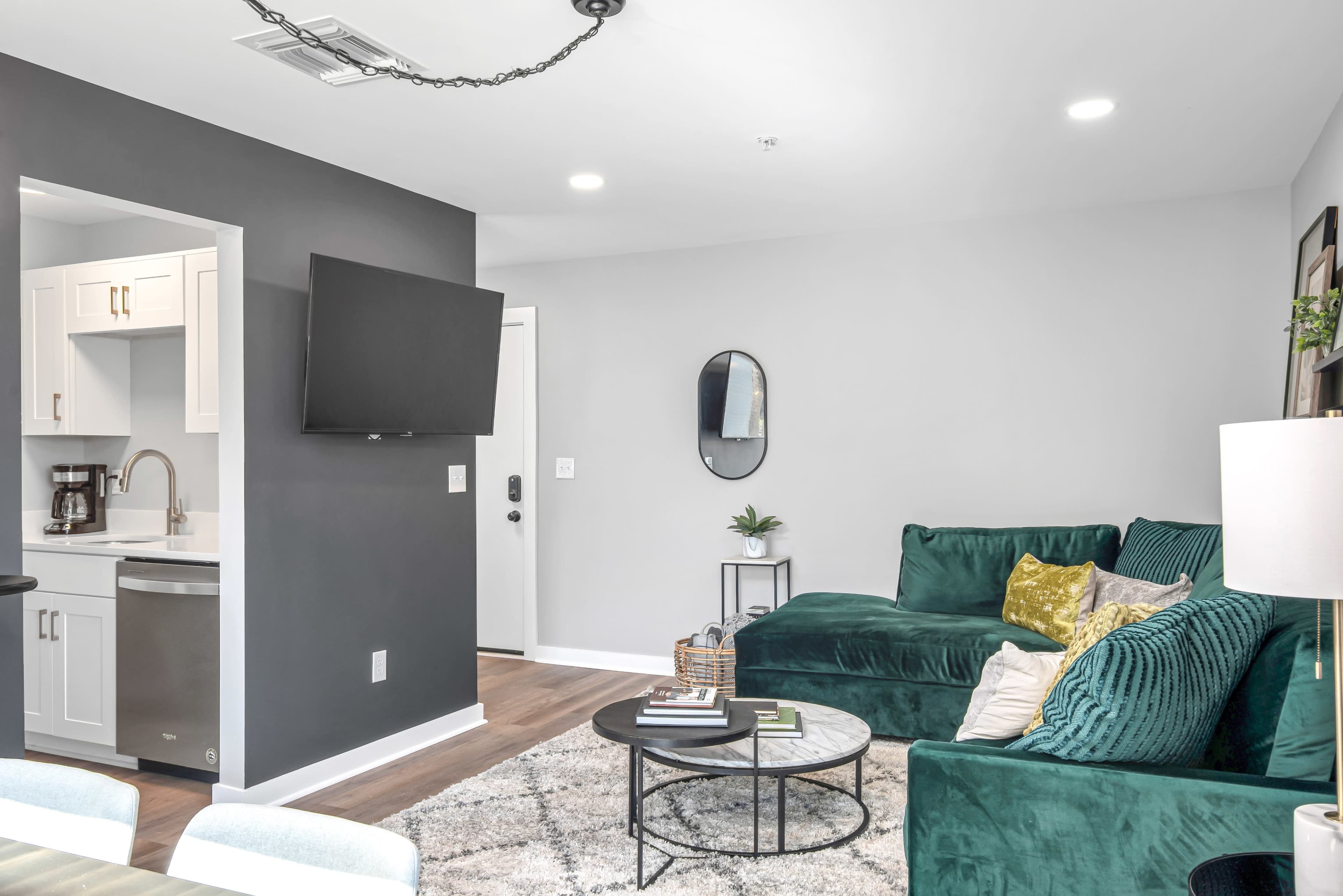 A modern living room featuring a green sectional sofa, a round coffee table on a patterned rug, and a kitchen area in the background with a television mounted on the wall.