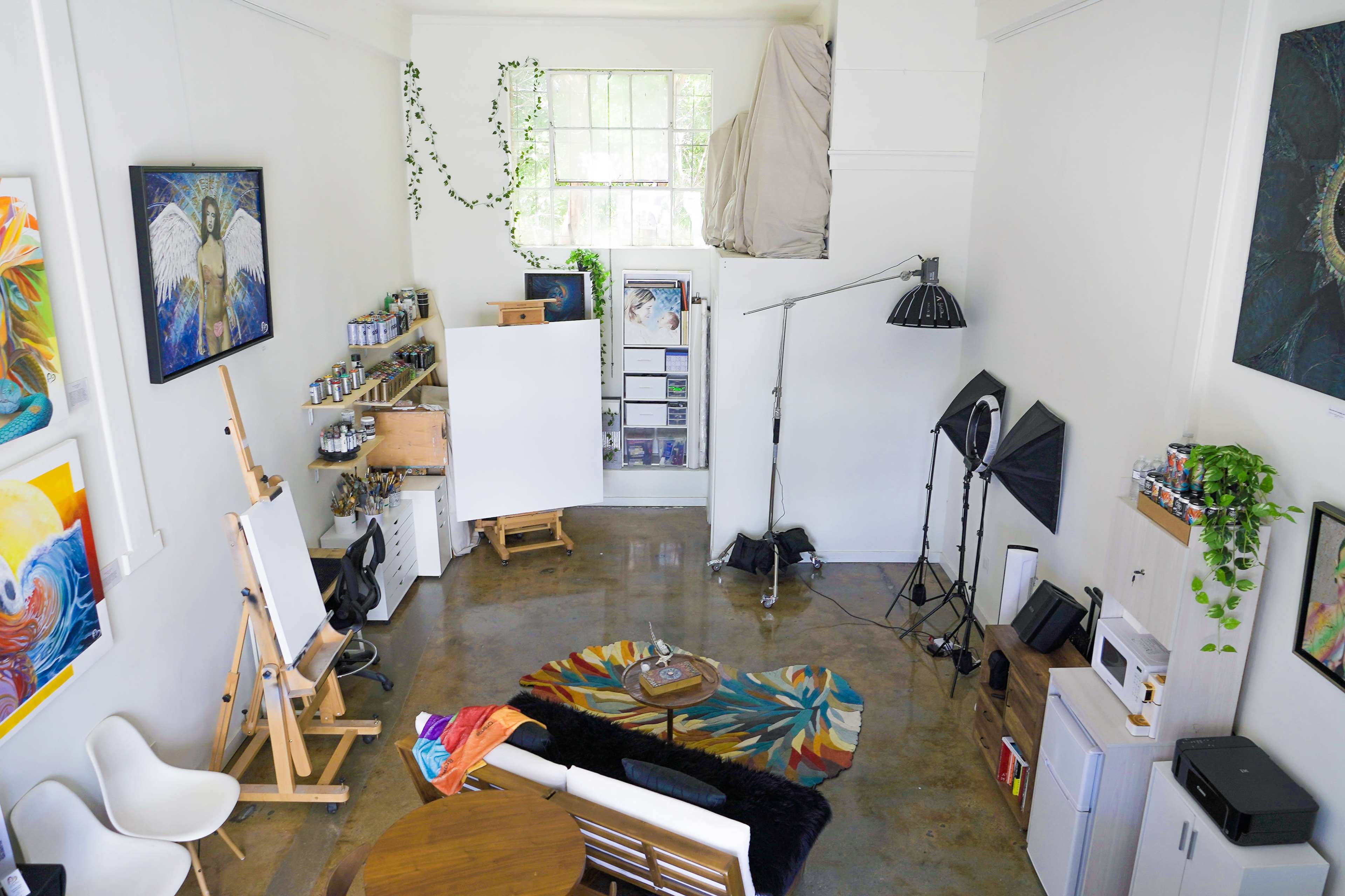 The image depicts an artist's studio featuring an easel, various art supplies, a seating area with a coffee table, and a backdrop for photography.