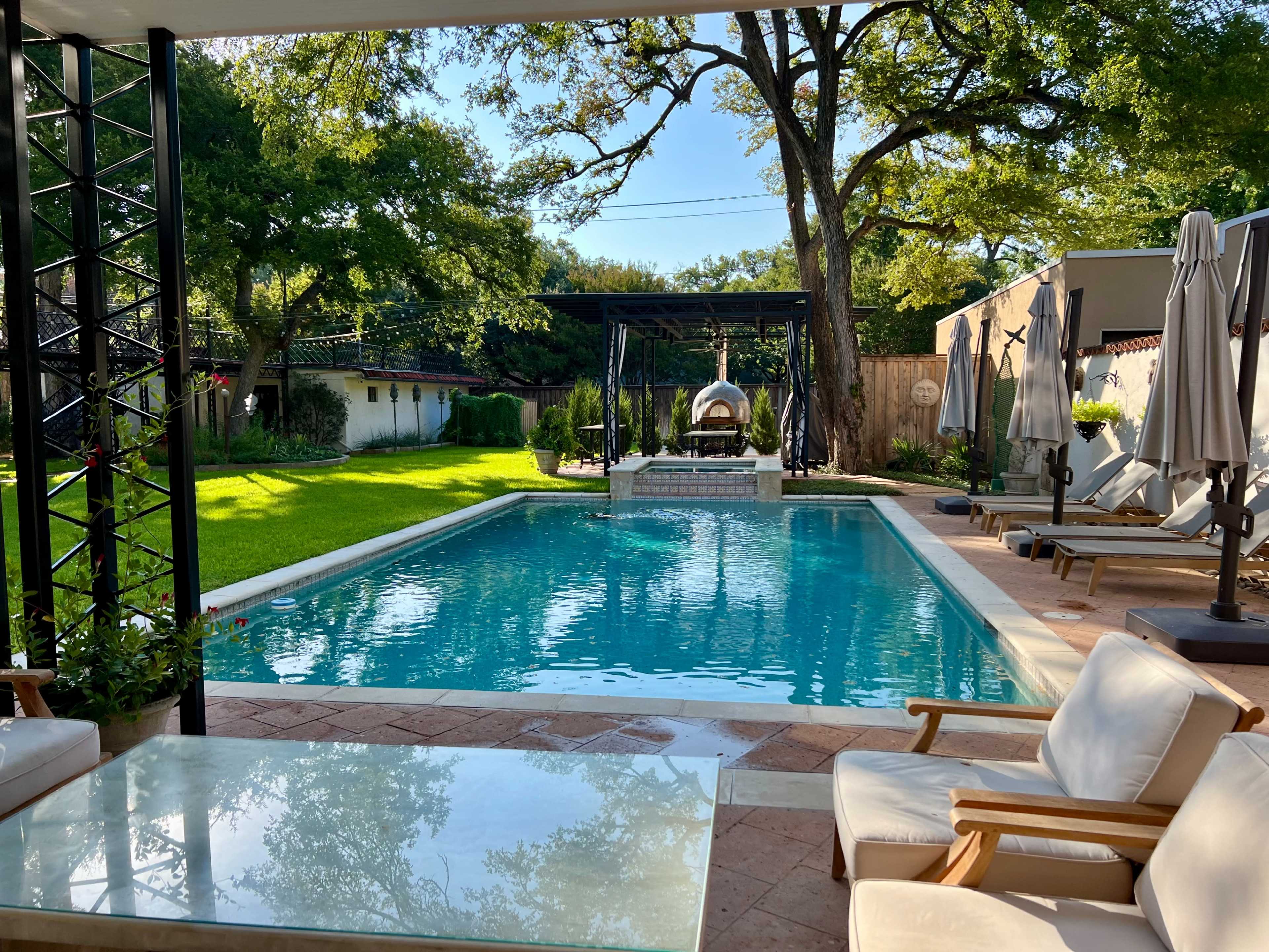 A clear swimming pool surrounded by lounge chairs and greenery, with shaded seating areas and a gazebo in the background.