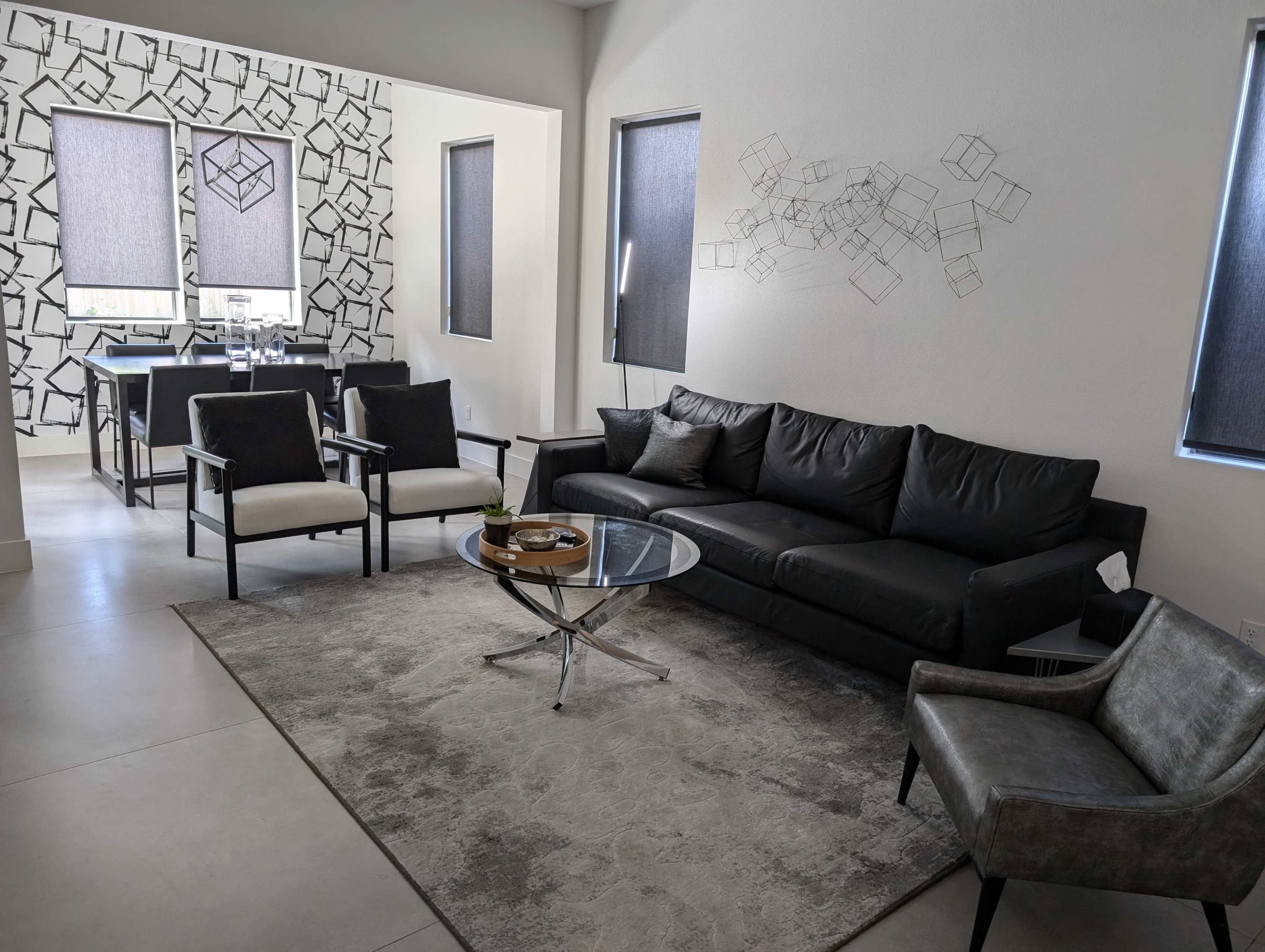 A modern living room featuring a black sofa, two armchairs, a glass coffee table, and a dining area in the background with a geometric wall design.