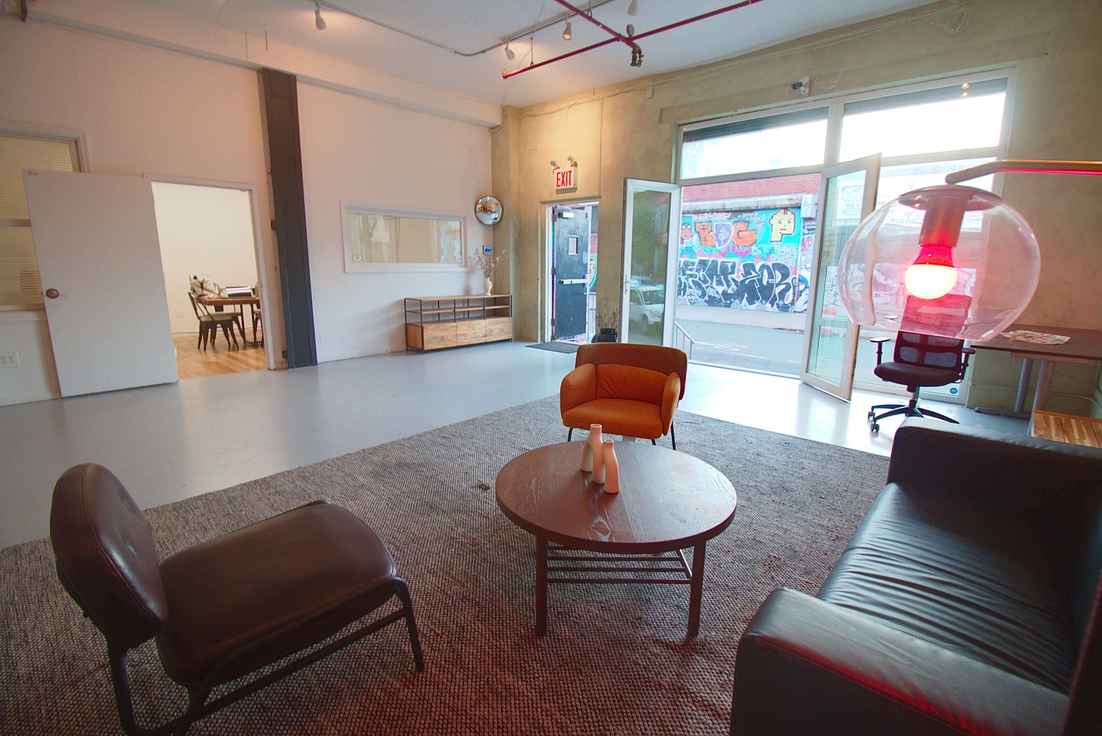 The image shows a modern interior space featuring a seating area with two chairs and a round table, large windows open to a mural-covered exterior wall, and a dining area in the background.