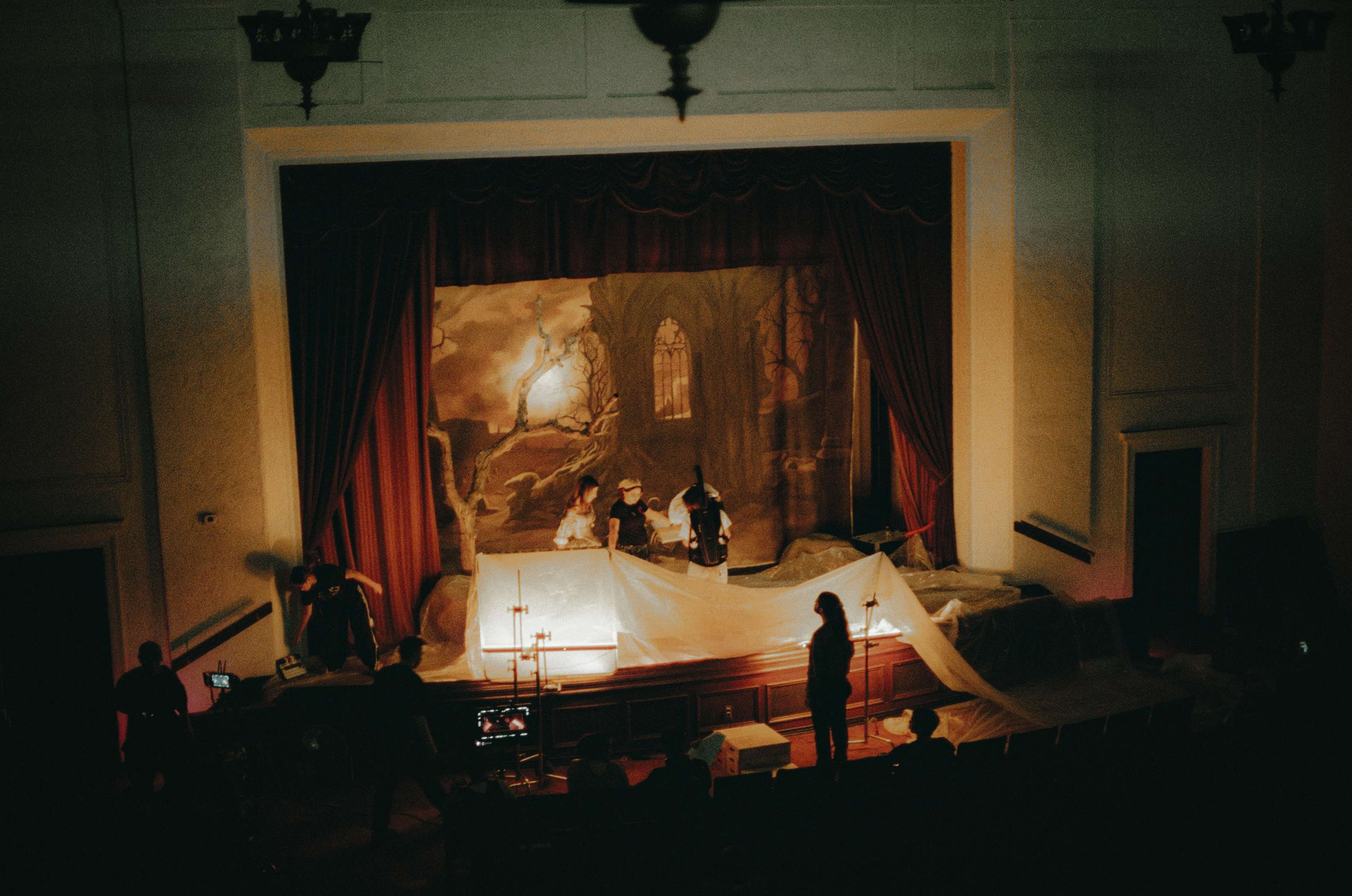 A group of people prepares a stage with a painted backdrop, draped in white fabric, in a theater setting.
