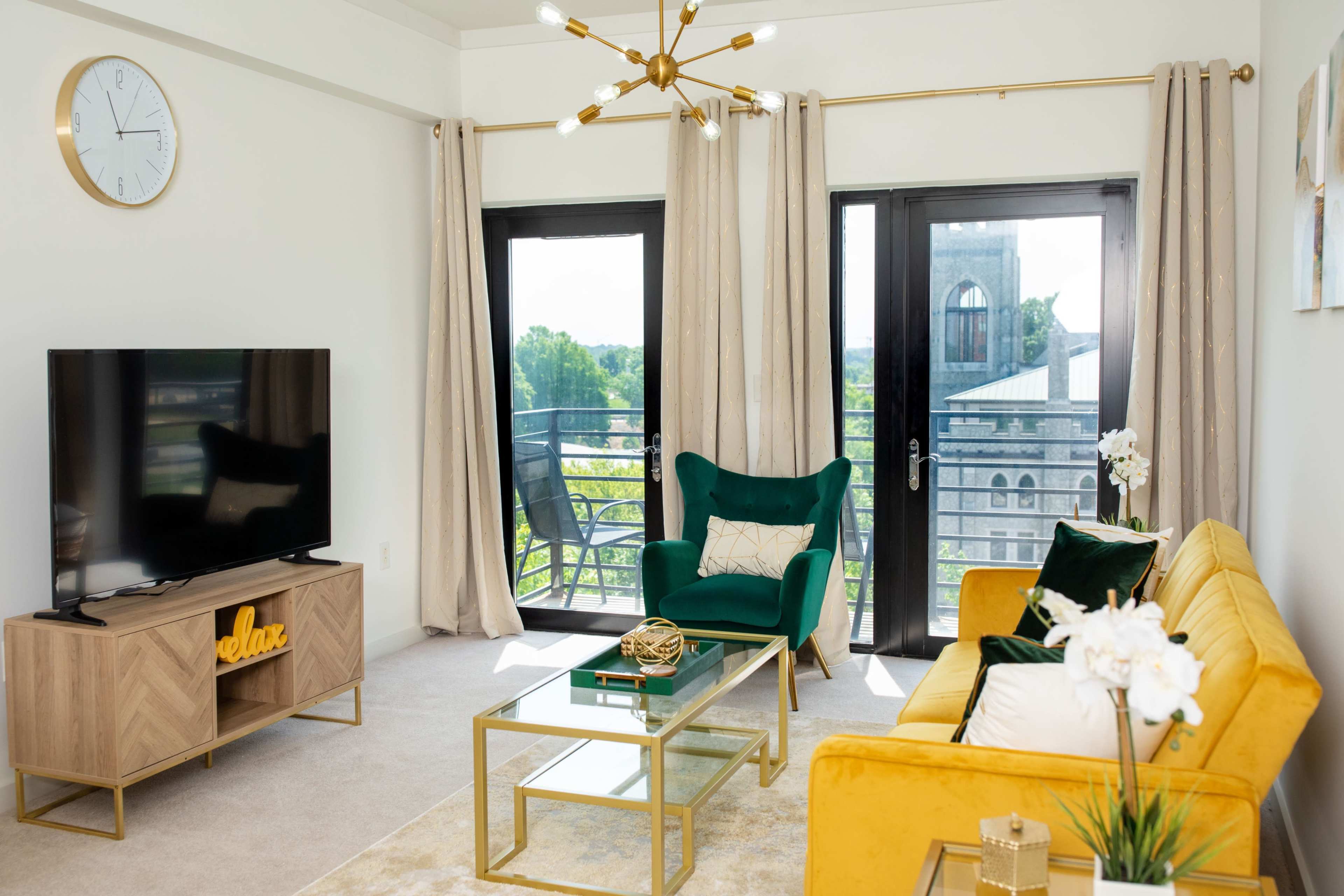 The image shows a modern living room featuring a green chair, a yellow sofa, a glass coffee table, and a television on a wooden stand, with large windows overlooking a balcony.