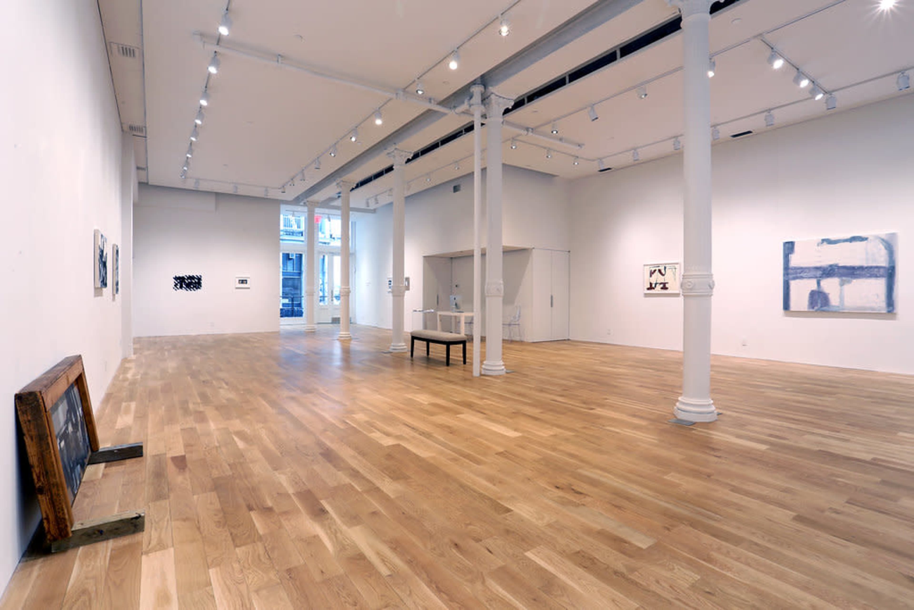 A spacious art gallery features polished wooden floors, tall white walls, and several pieces of artwork displayed among structural columns.