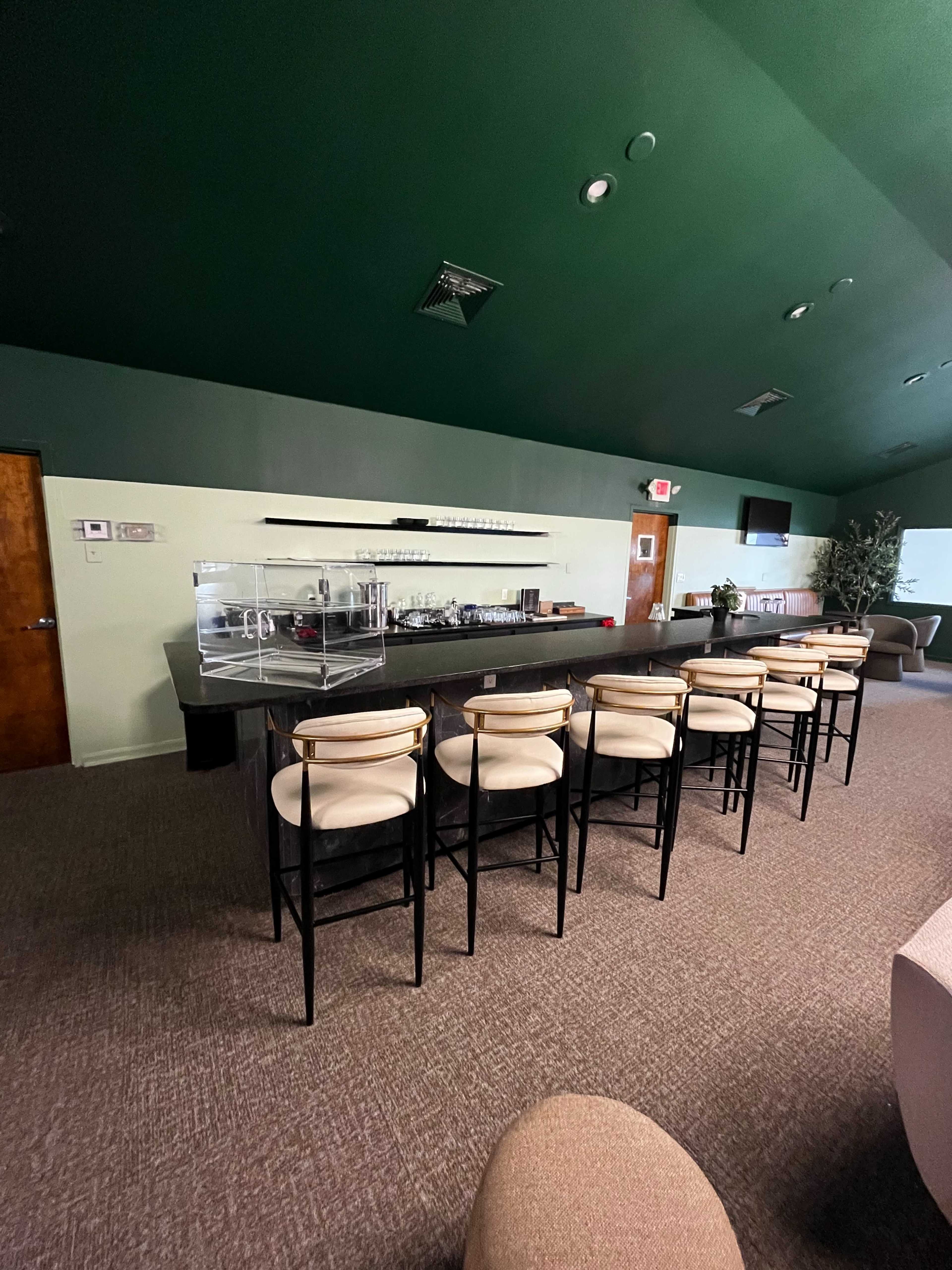 The image shows a modern lounge area featuring a bar with tall black stools and a glass display case on a green wall backdrop.