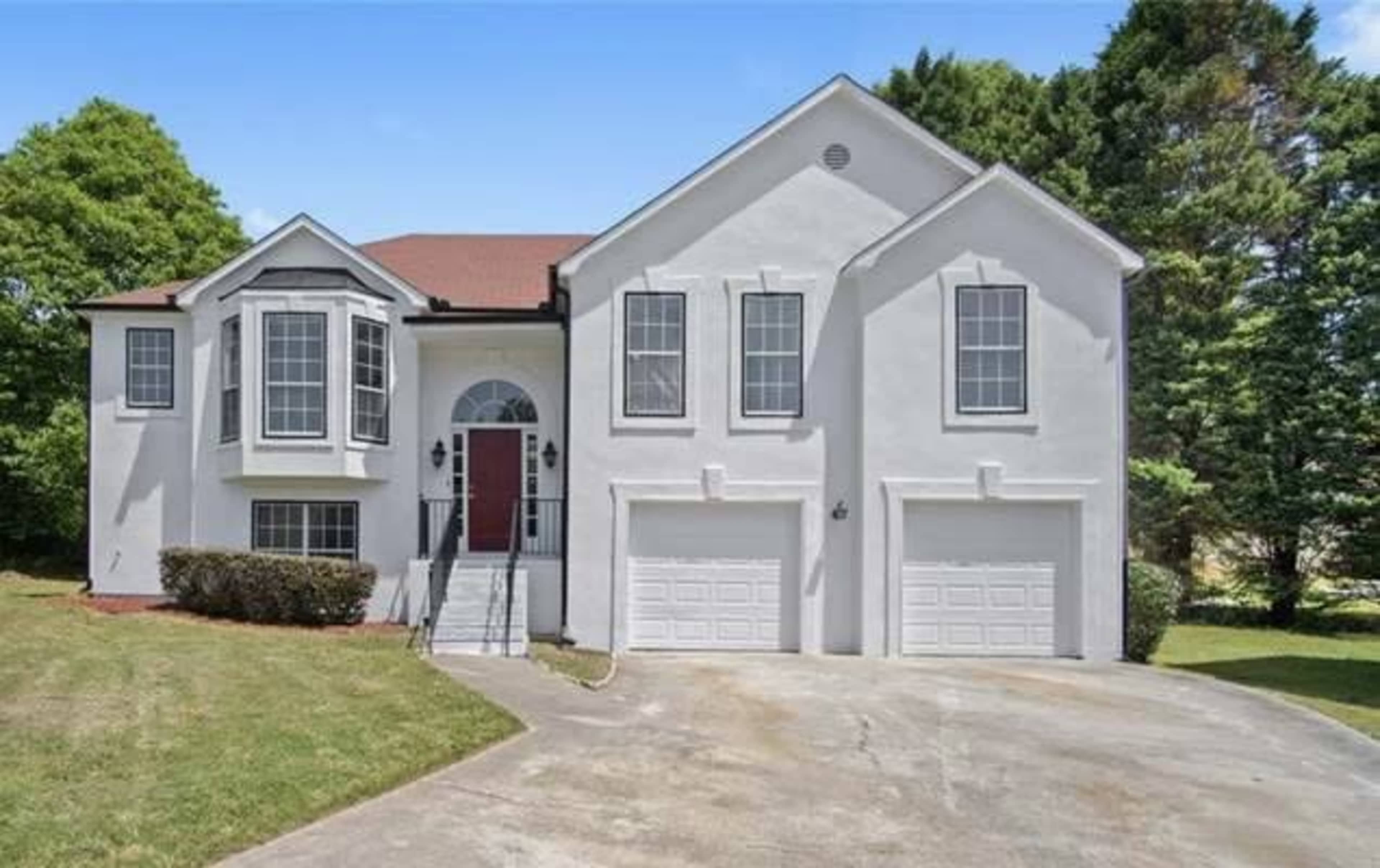 The image shows a two-story, modern white house with a red front door and two attached garages, set on a green lawn under a clear blue sky.