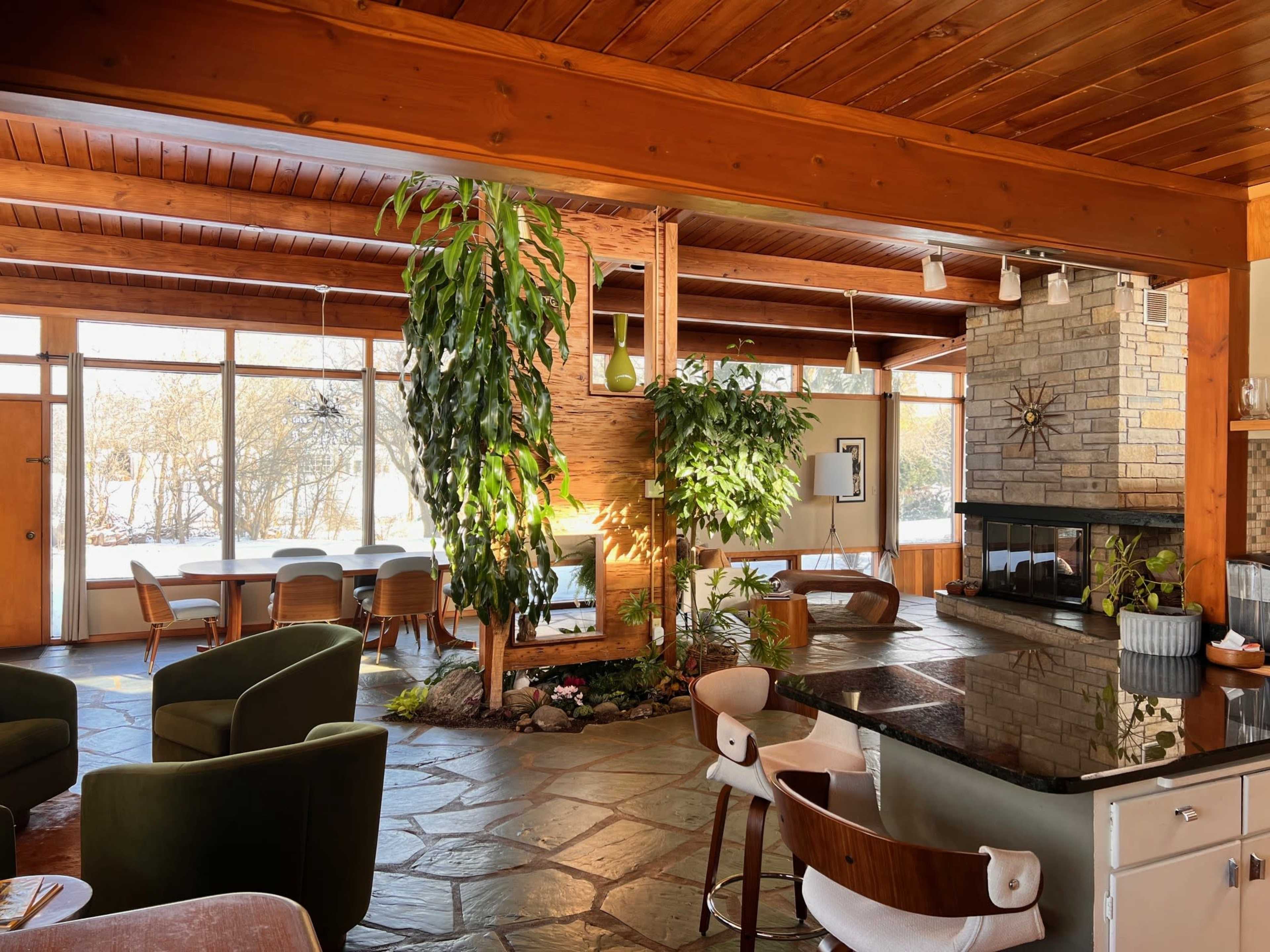 The image shows a spacious interior with a stone fireplace, large windows, plants, and a kitchen area featuring a dark countertop.