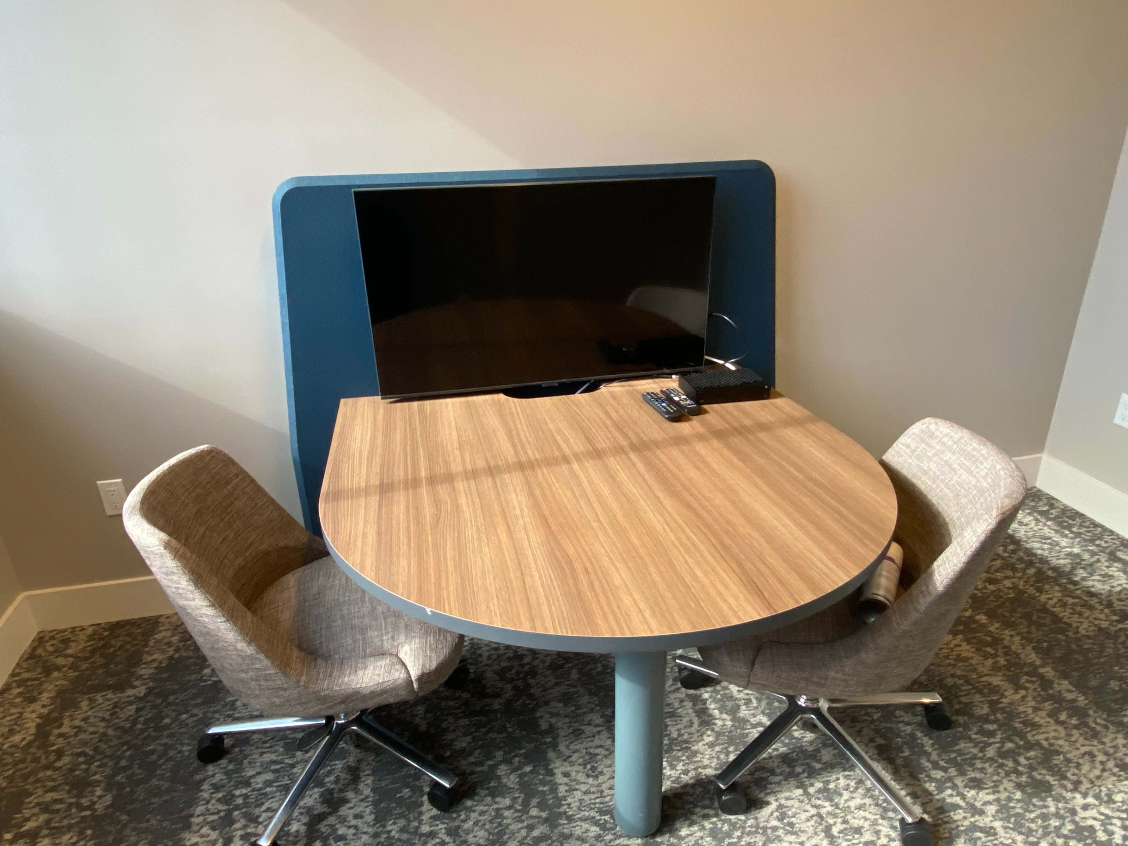 A round wooden table with two chairs and a wall-mounted television is set against a neutral wall.