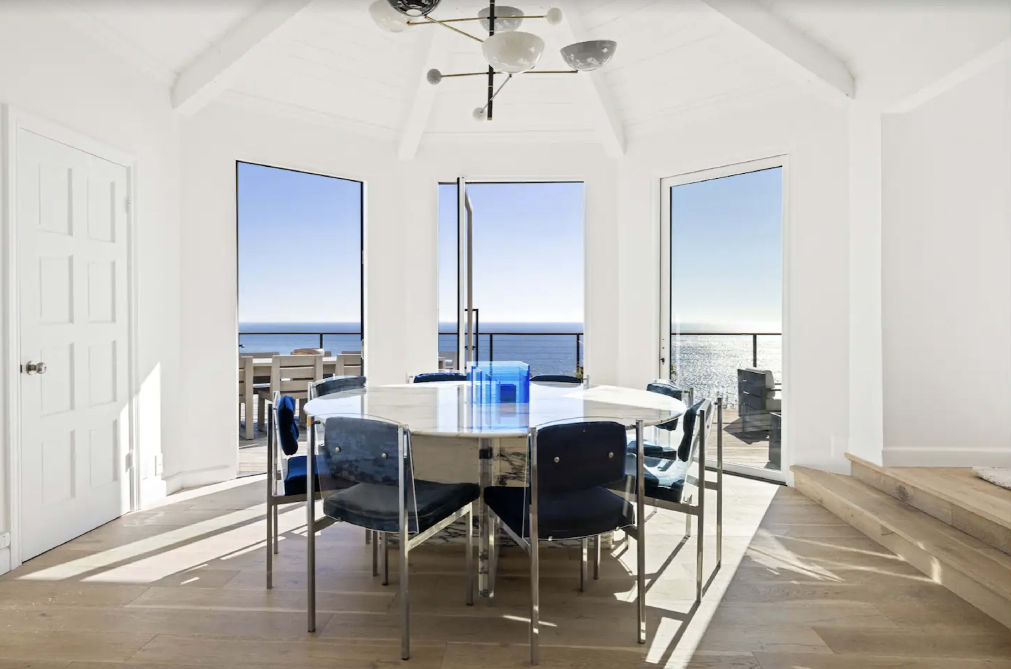 A modern dining area with a circular table surrounded by clear chairs, featuring large windows that offer a view of the ocean.