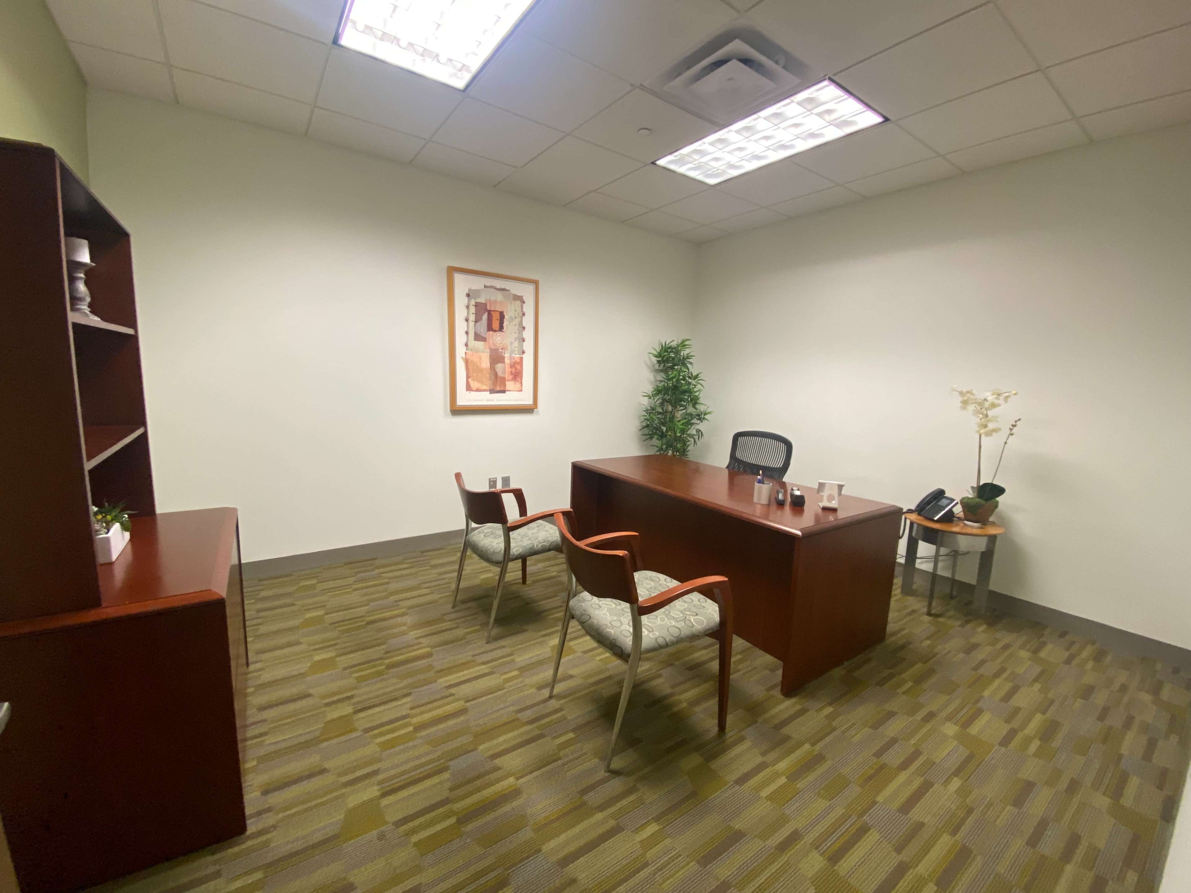 An empty office space features a wooden desk, two chairs, a bookshelf, and a plant against a neutral wall.