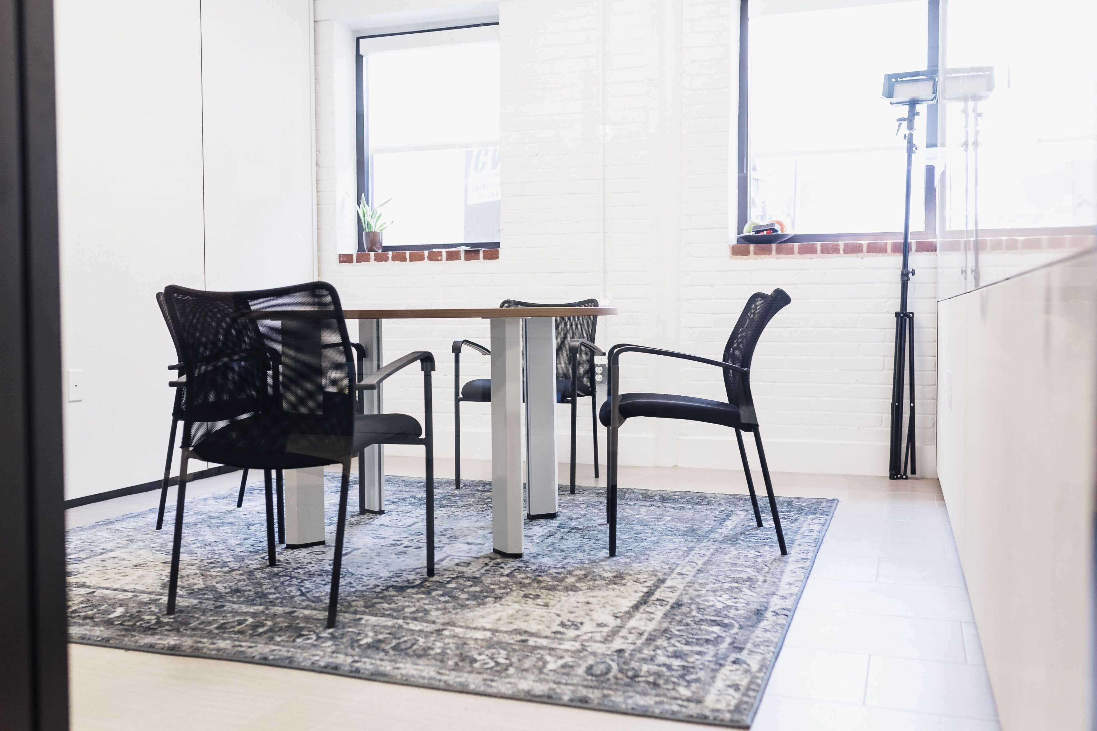 A small meeting room with a wooden table surrounded by four black chairs on a patterned rug, with large windows letting in natural light.