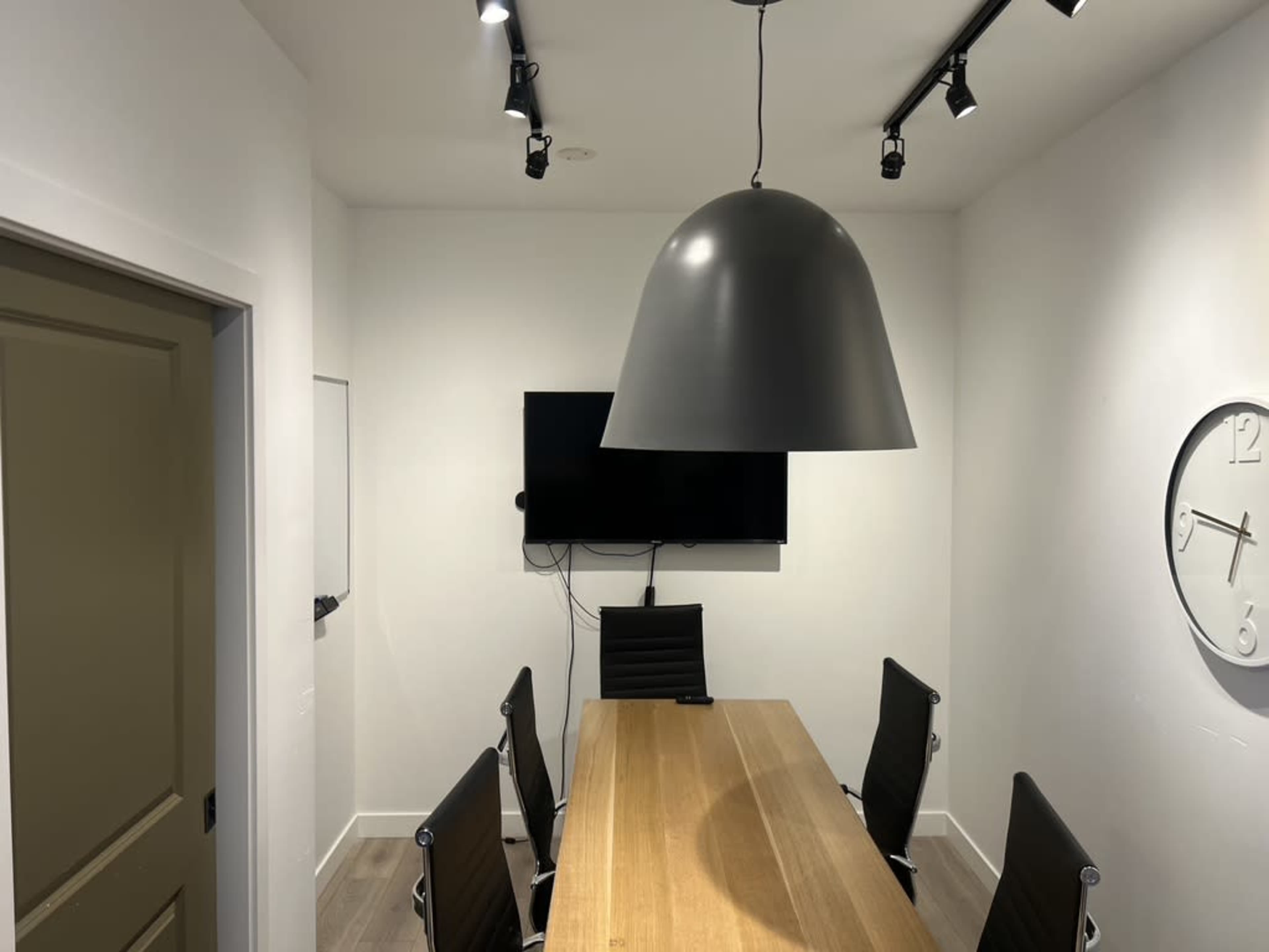 A conference room features a large wooden table surrounded by black chairs, a flat-screen TV mounted on the wall, and a large pendant light hanging from the ceiling.