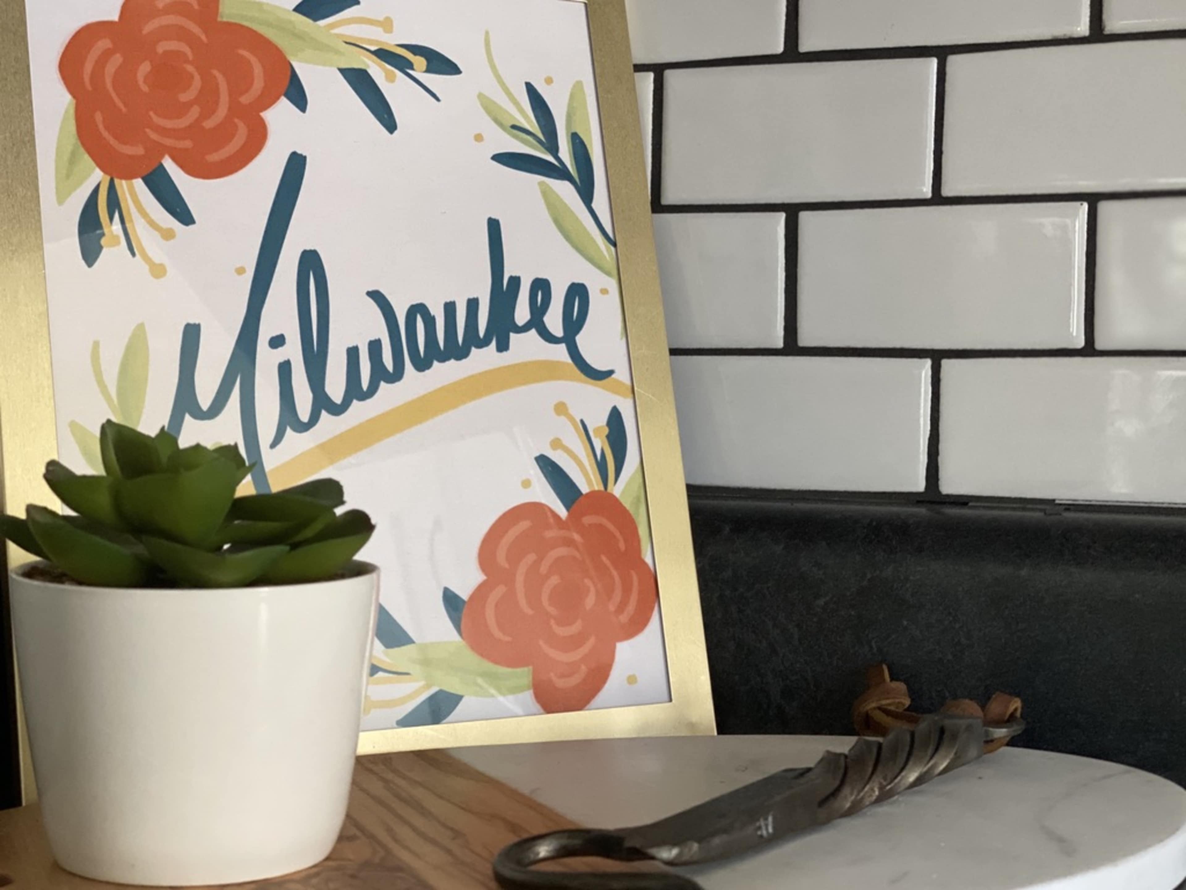A framed decorative artwork with "Milwaukee" written in cursive is displayed next to a small potted succulent on a marble surface, alongside a vintage metal object.