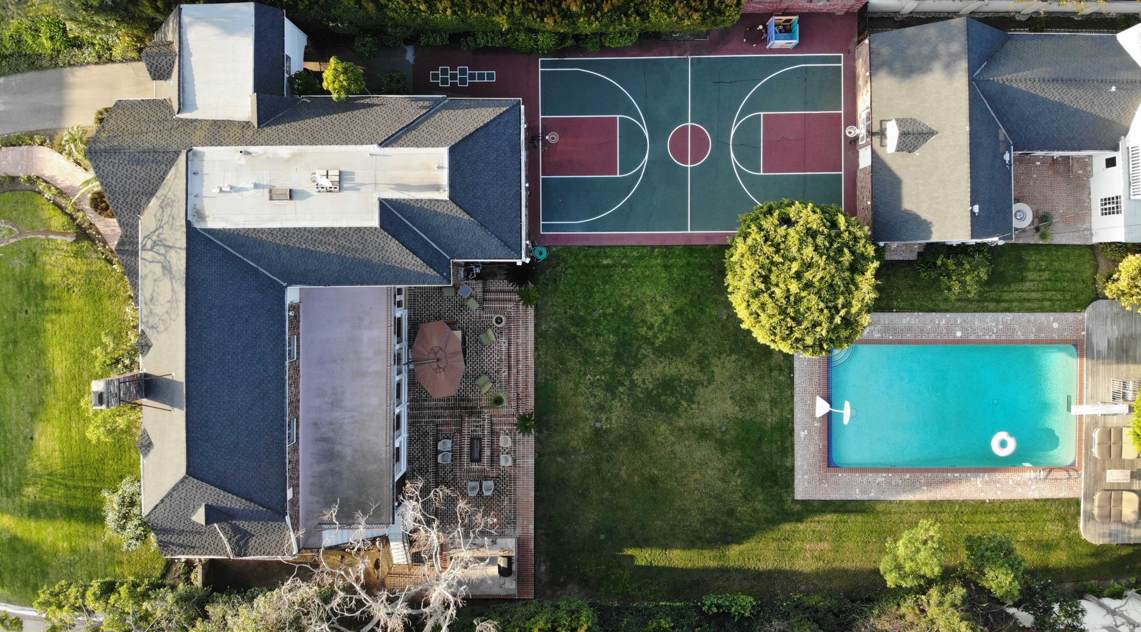 An aerial view of a residential property featuring a house, a basketball court, and a swimming pool surrounded by landscaped gardens.