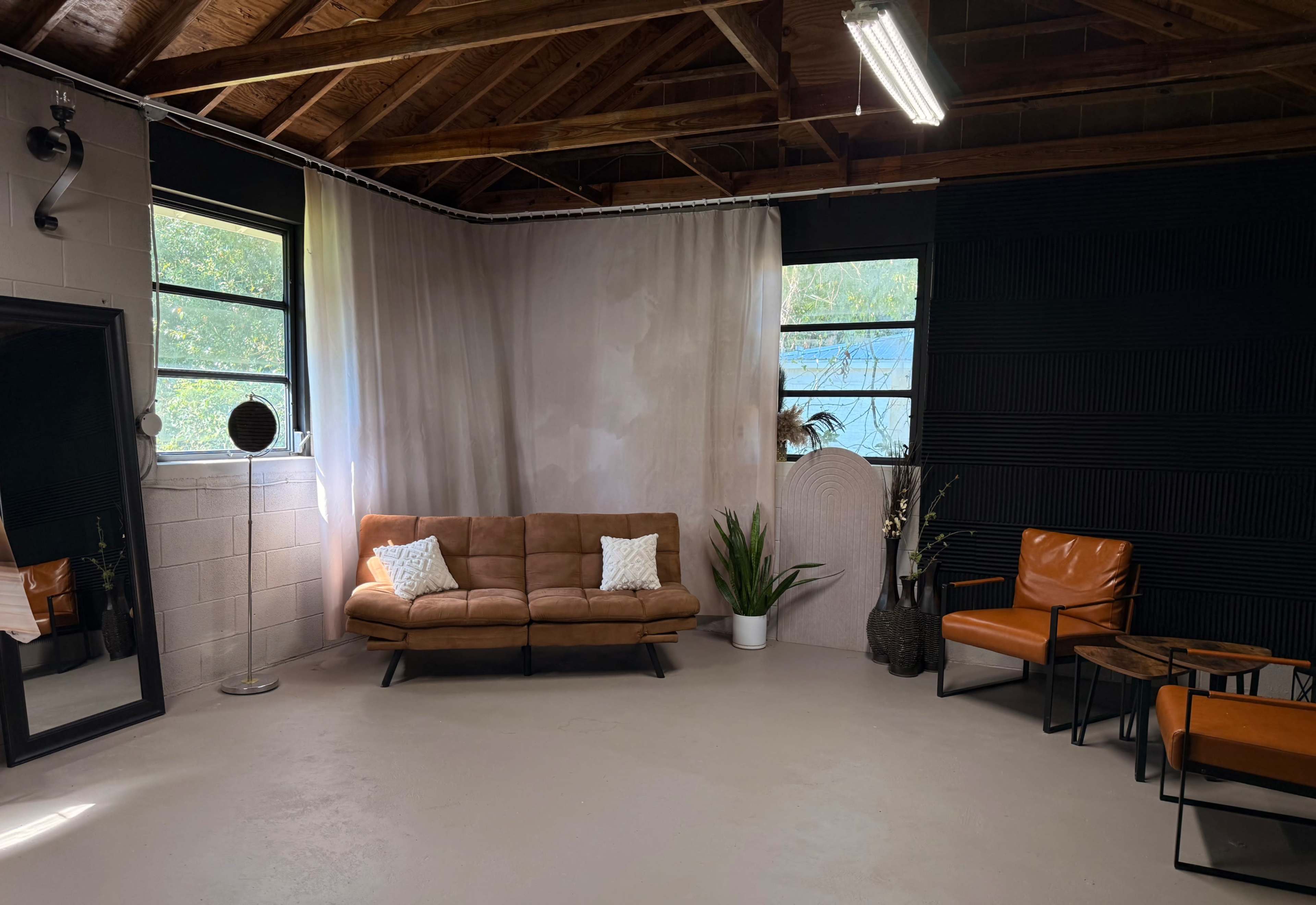 The image shows a spacious, uncluttered room with a brown sofa, orange armchairs, a large mirror, and large windows that allow natural light to enter.