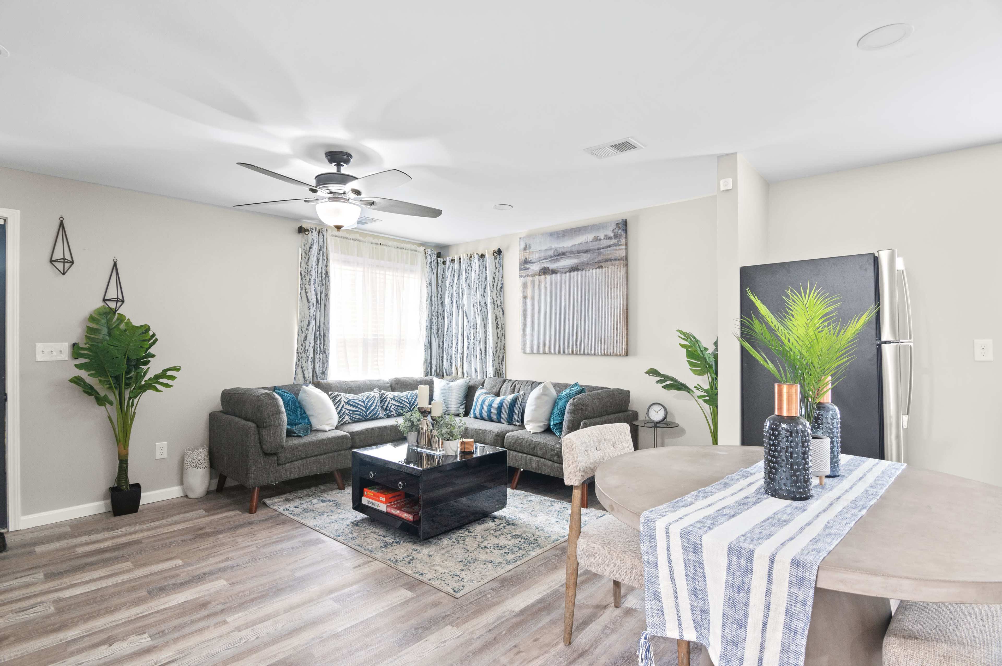 The image depicts a modern living room with two gray sofas, a glass coffee table, and a dining area featuring a round table set for four.
