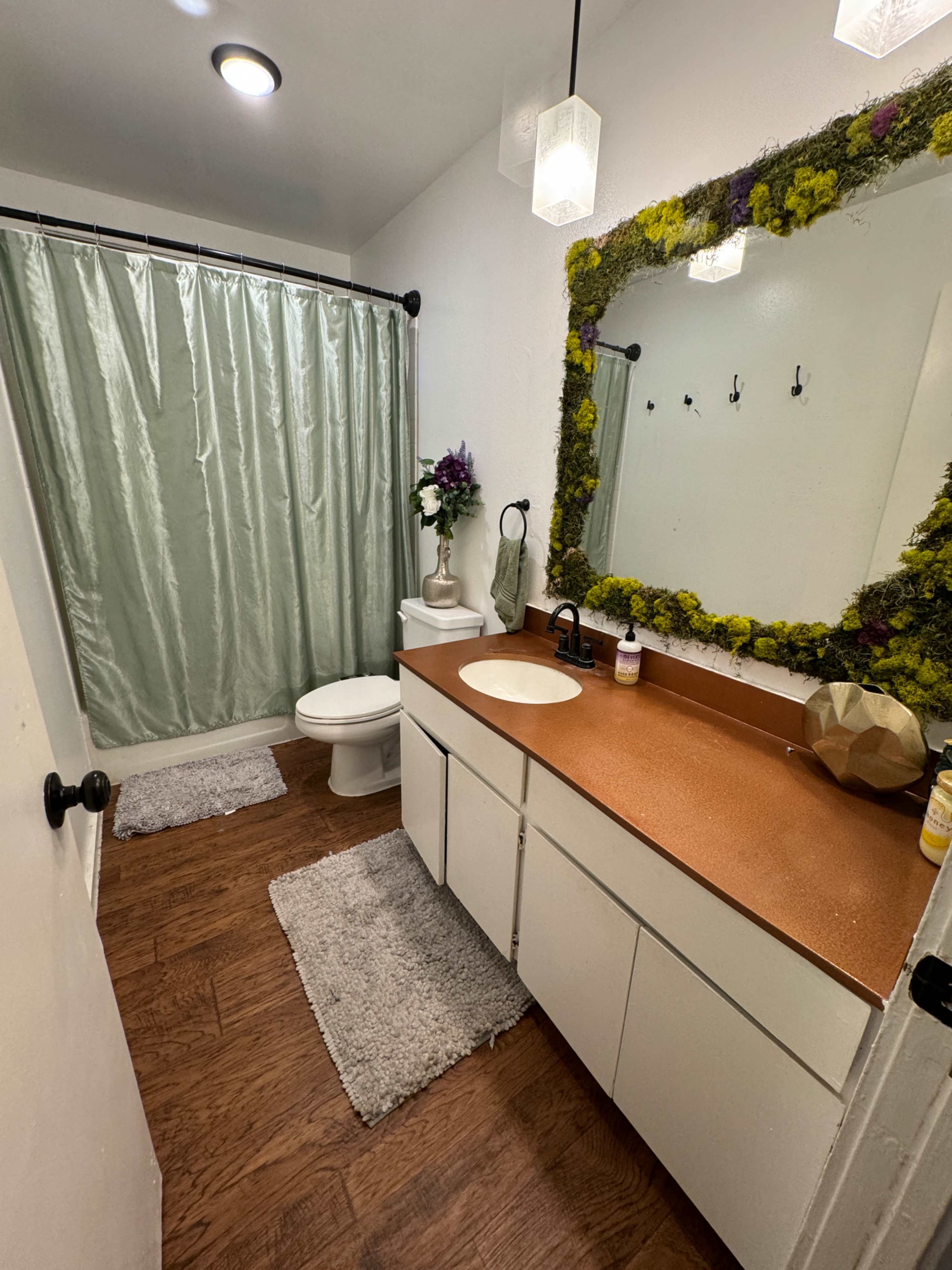 The bathroom features a toilet, a sink with a countertop, a shower with a green curtain, and a mirror framed with decorative moss.
