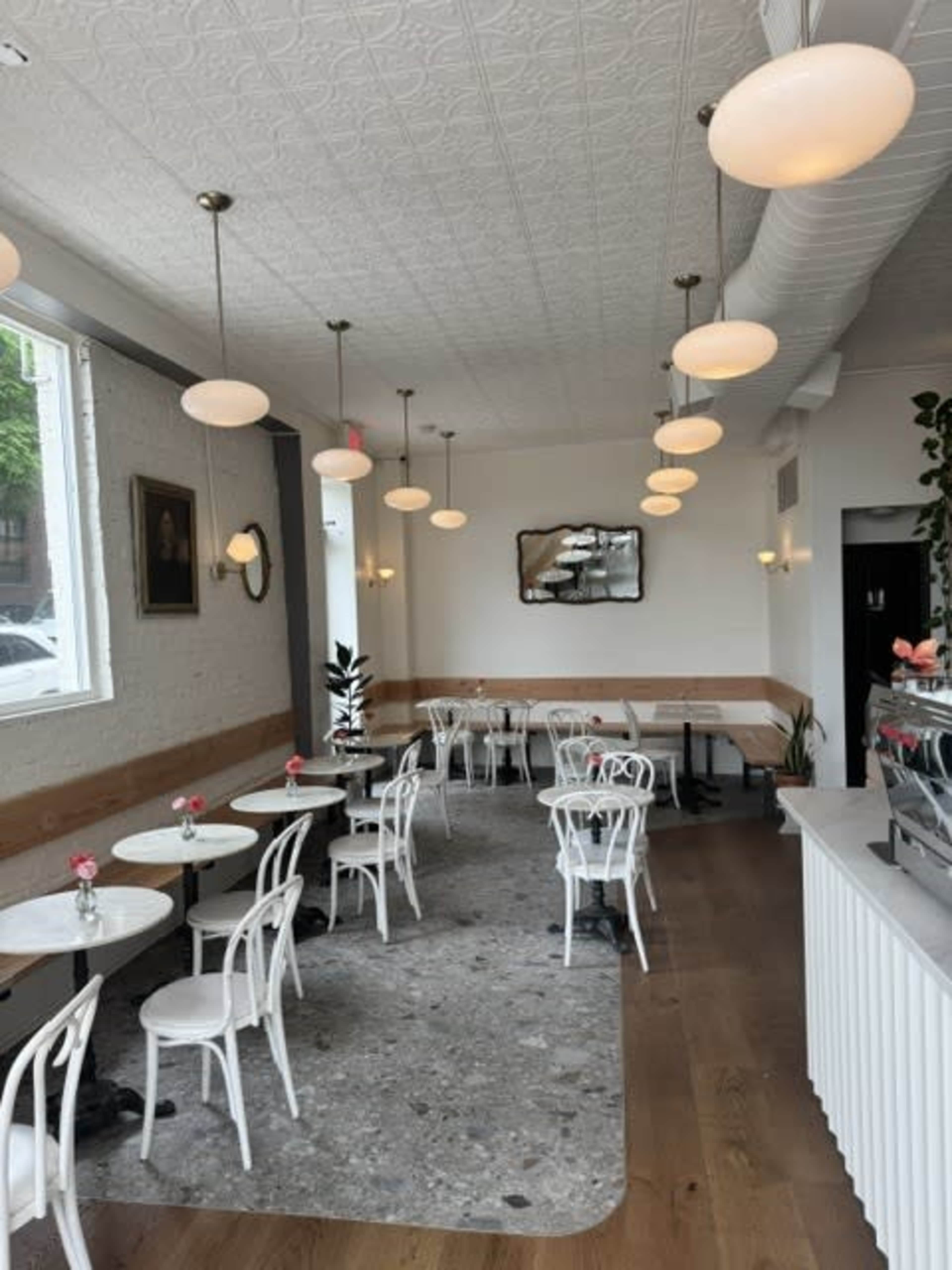 The image shows a light-filled café with white chairs and tables, featuring pendant lights and a simple décor.