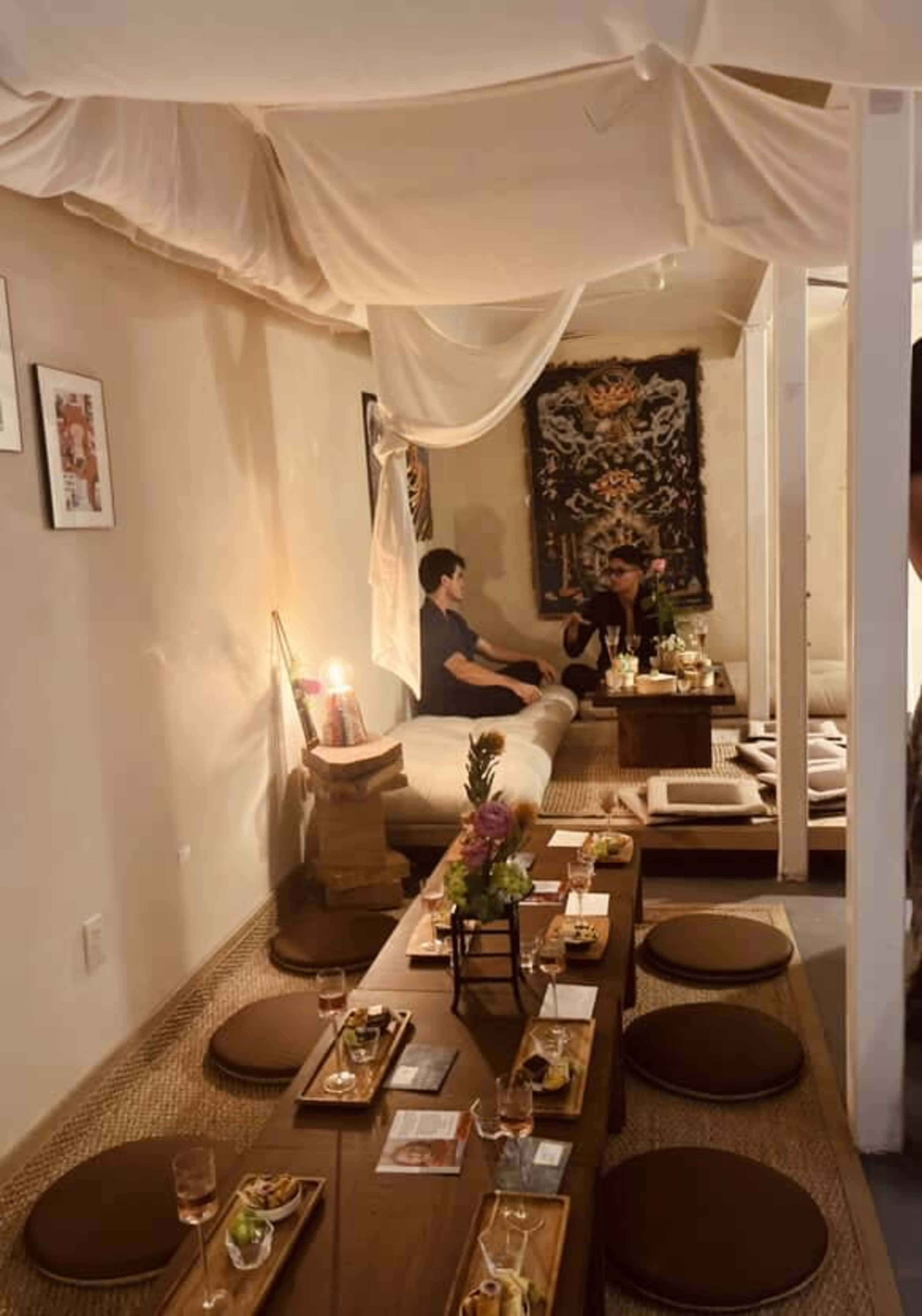 The image shows a serene dining area with low tables and cushions, featuring two people seated and enjoying their meal in a softly lit, draped environment.