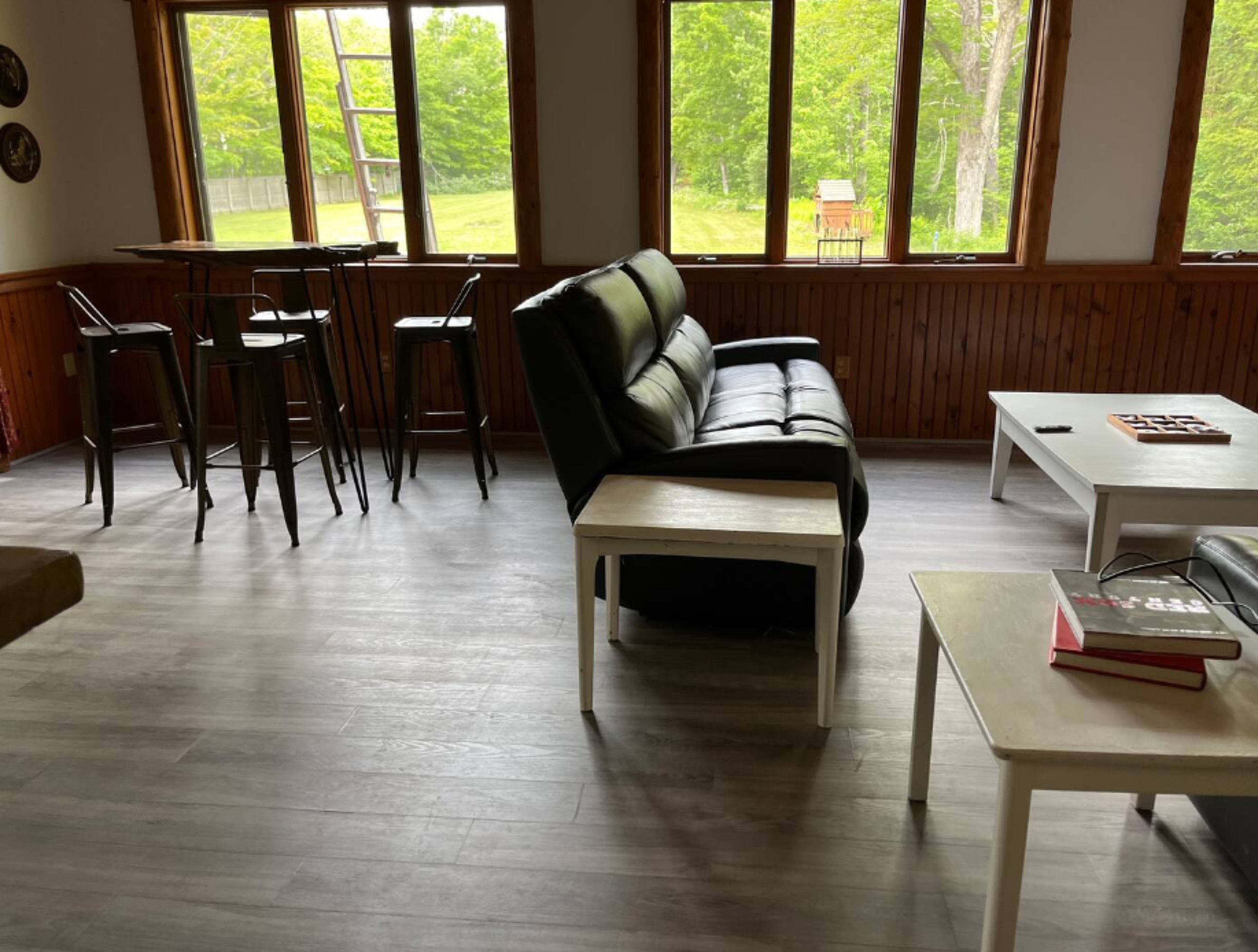 A spacious living area with a black leather sofa, several white coffee tables, and tall bar stools near large windows overlooking a green landscape.