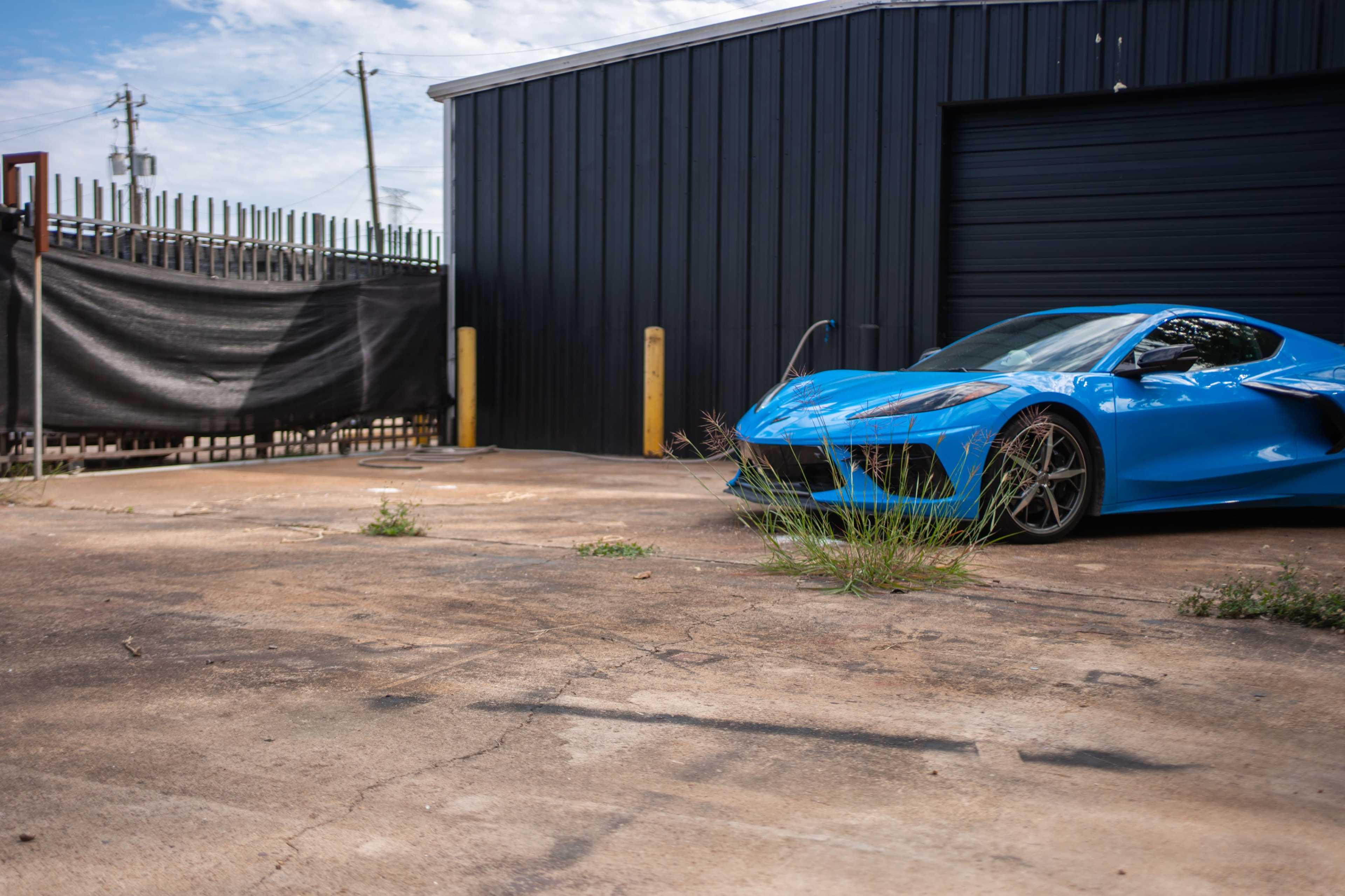 A blue sports car is parked on a concrete surface next to a black building, with grass and small weeds growing through the cracks.