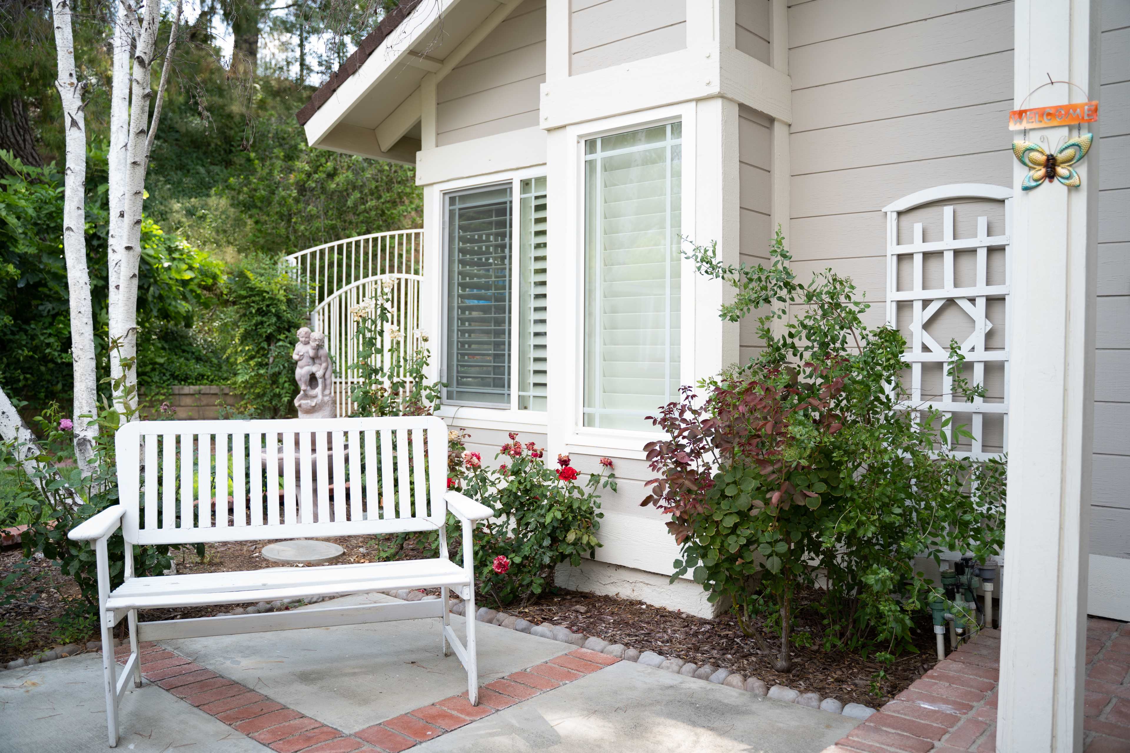 A white bench sits on a patio near a house with green plants and bushes, framed by a white trellis and decorative statue.