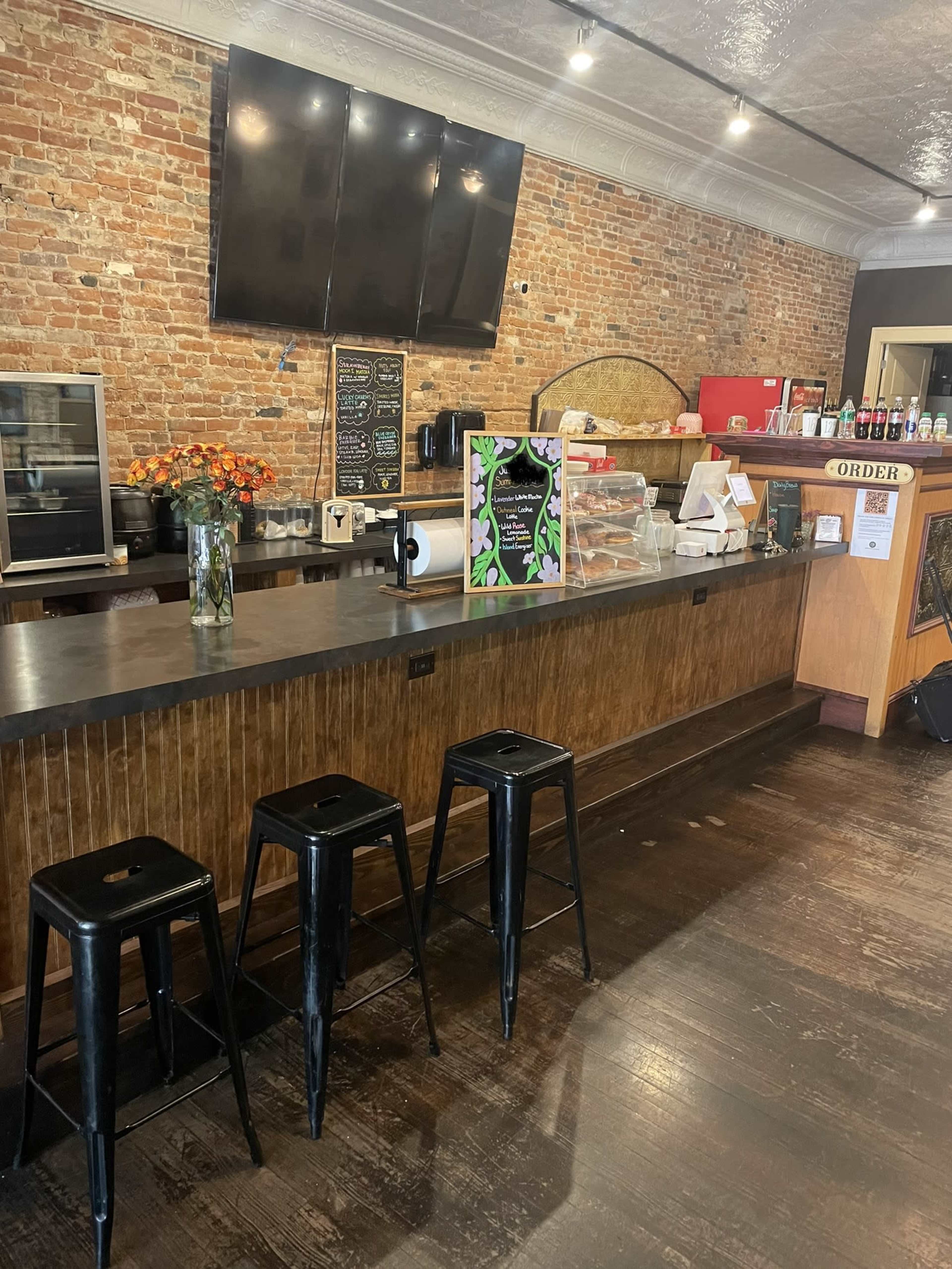 A rustic café interior featuring a wooden counter, three black stools, and exposed brick walls, with a flower arrangement on the counter and a television mounted on the wall.