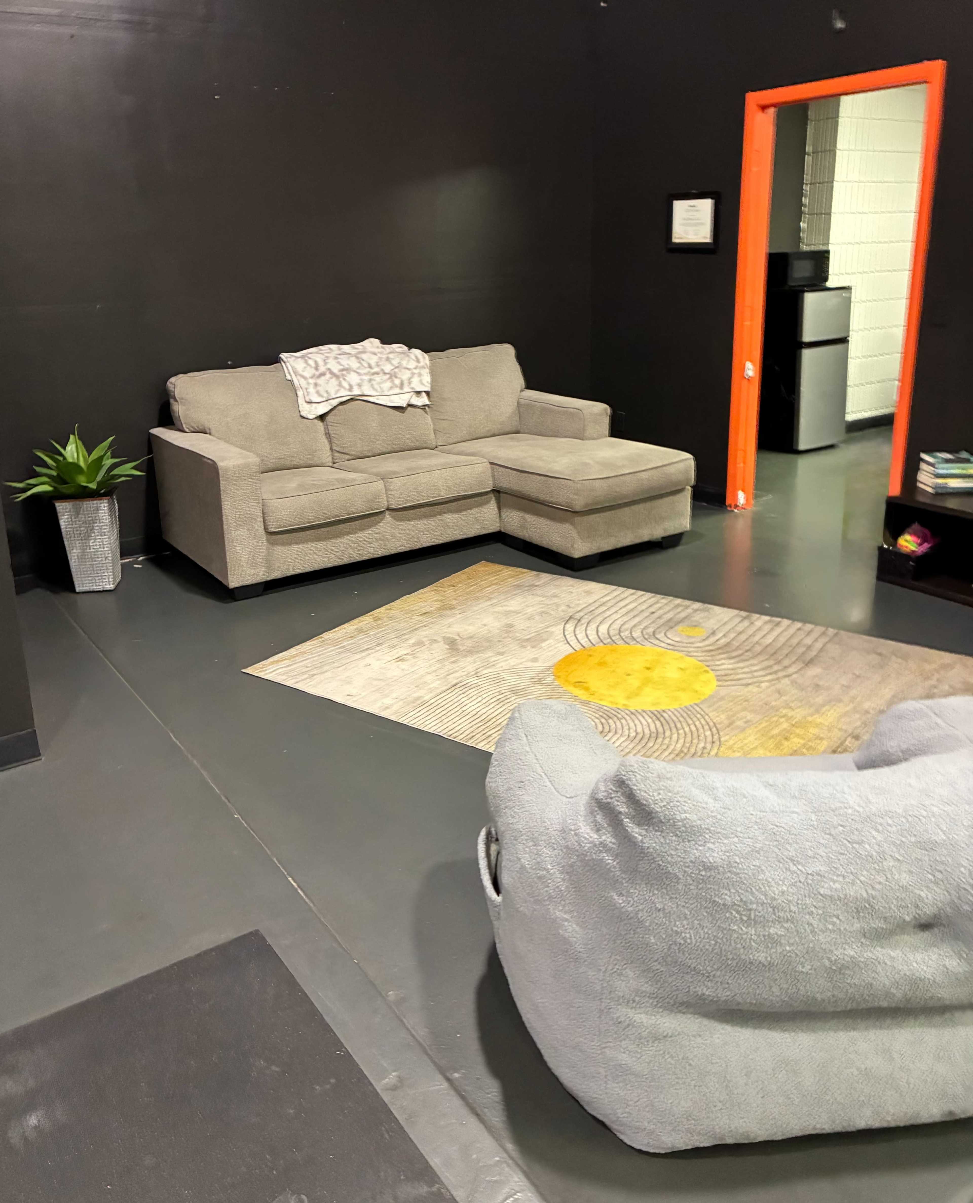 The image shows a seating area with a gray sectional couch and a light gray armchair, in a room with black walls and an orange door leading to a kitchen.