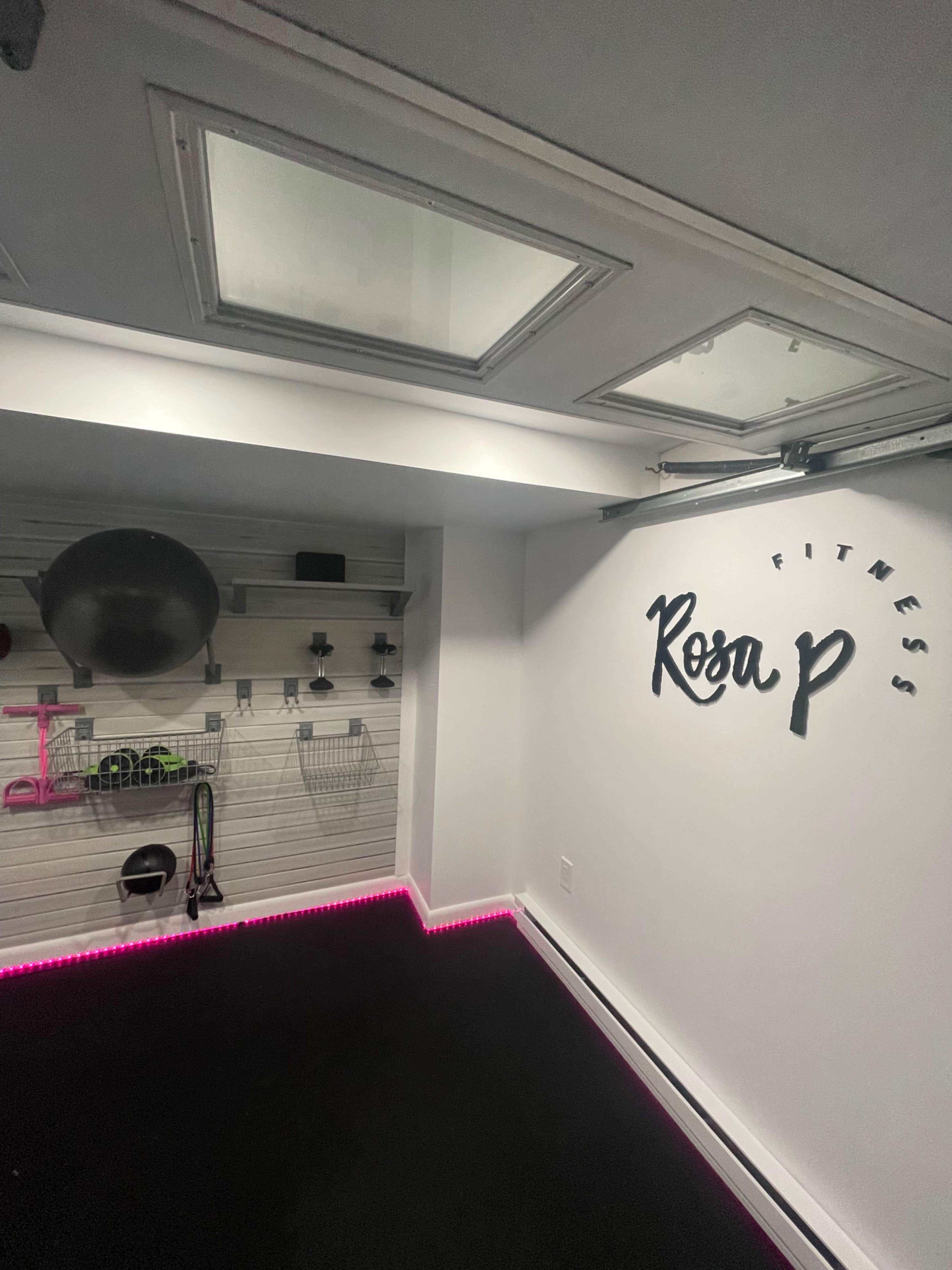 A modern fitness room with wall-mounted storage for exercise equipment and a lit logo reading "Rosa P Fitness" on the wall.