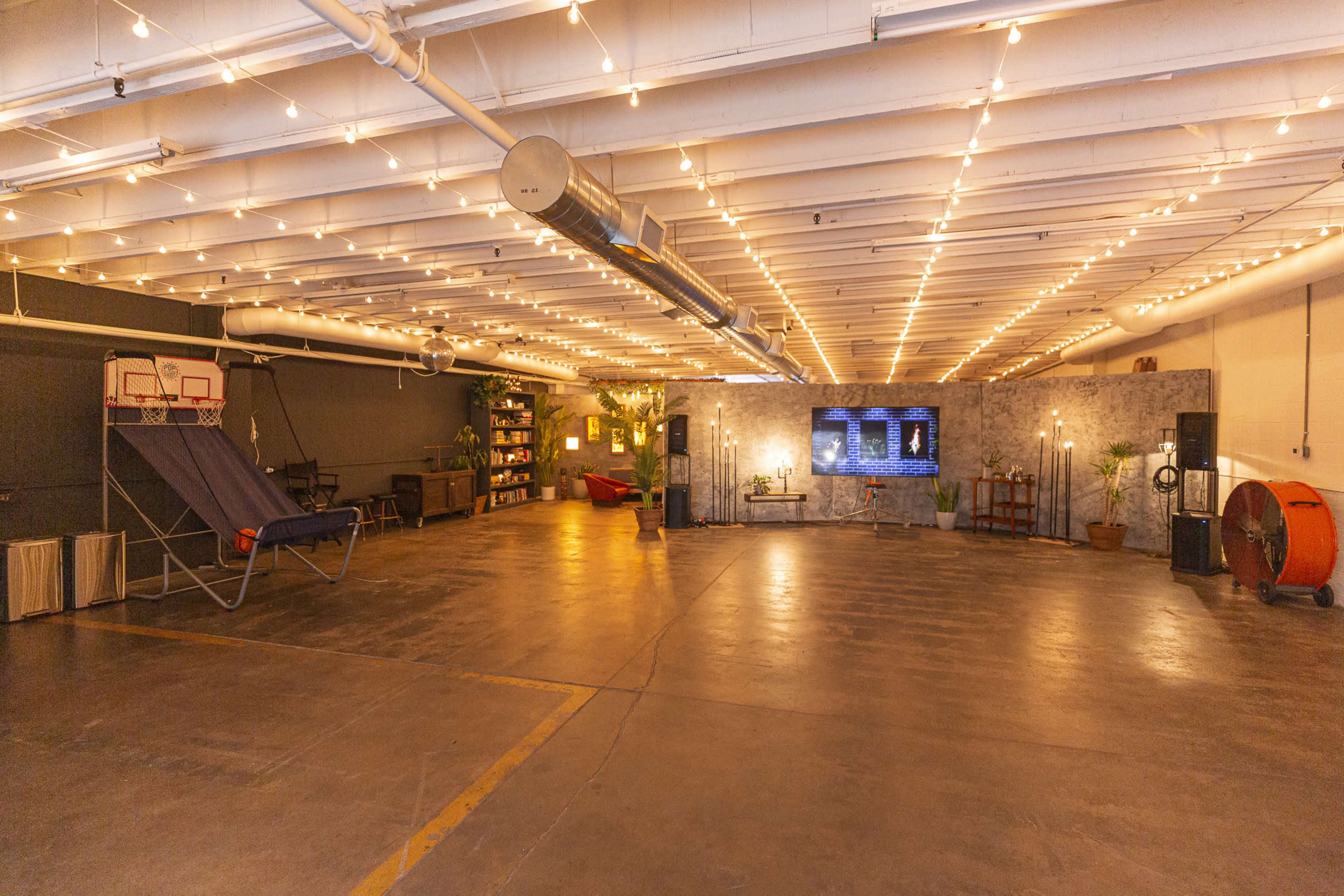 The image shows a spacious indoor area with string lights, a basketball hoop, seating, shelves, and a large screen on the wall.