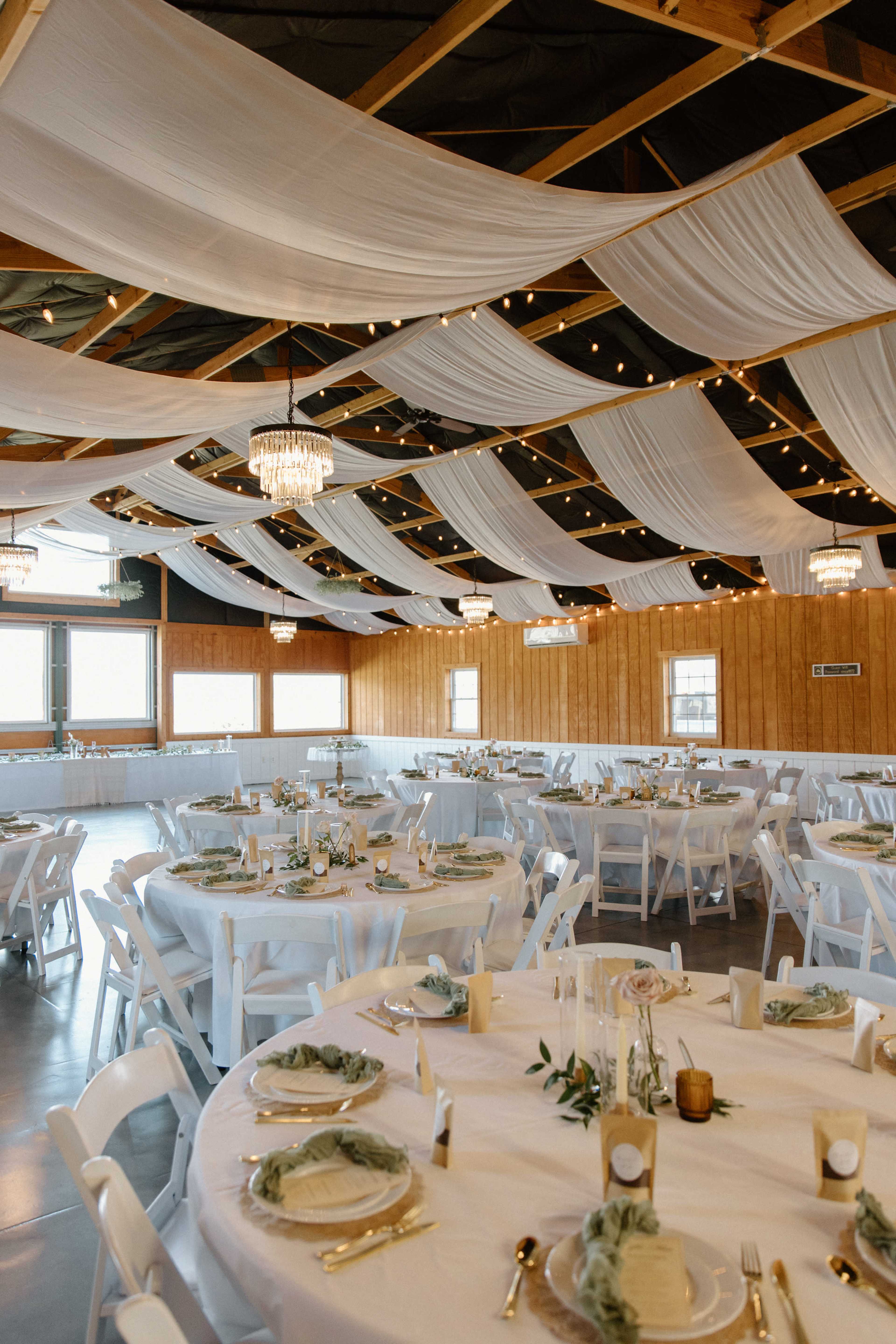 The image shows a decorated event space with white drapes and string lights overhead, featuring round tables set with elegant tableware and floral centerpieces.