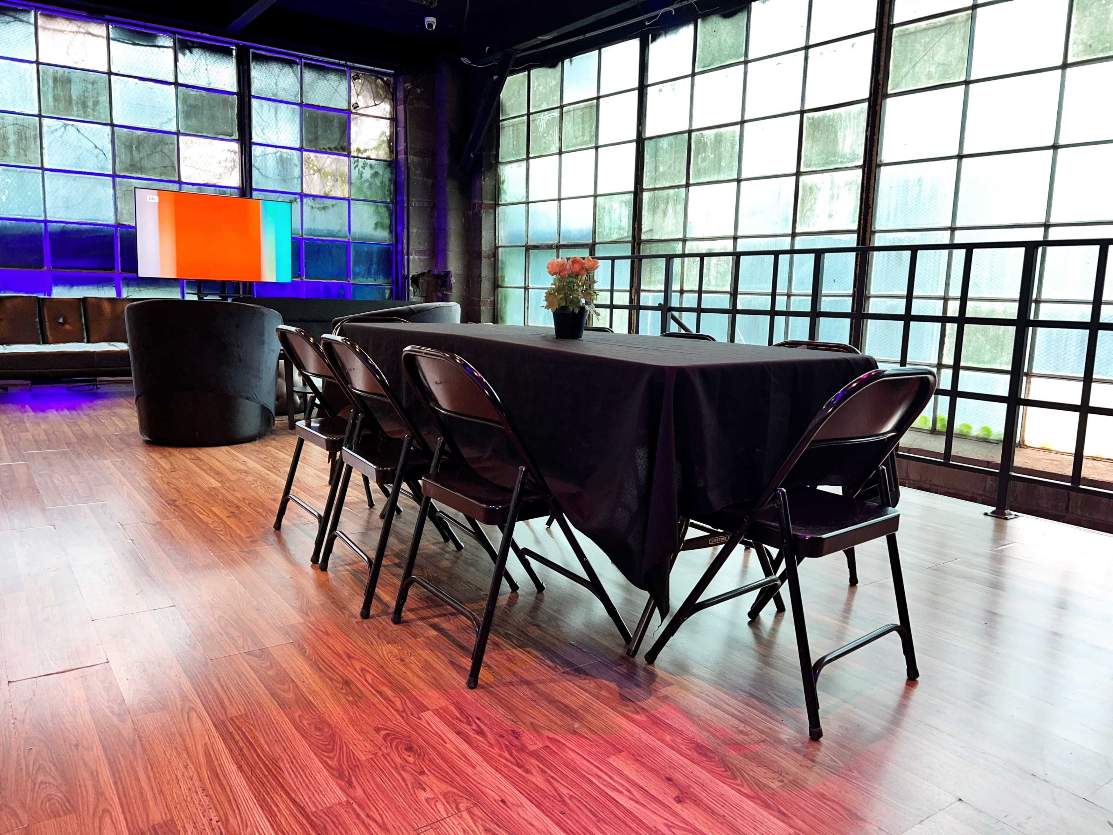 A long black table with six folding chairs is set up in a modern room with large windows and a television screen in the background.