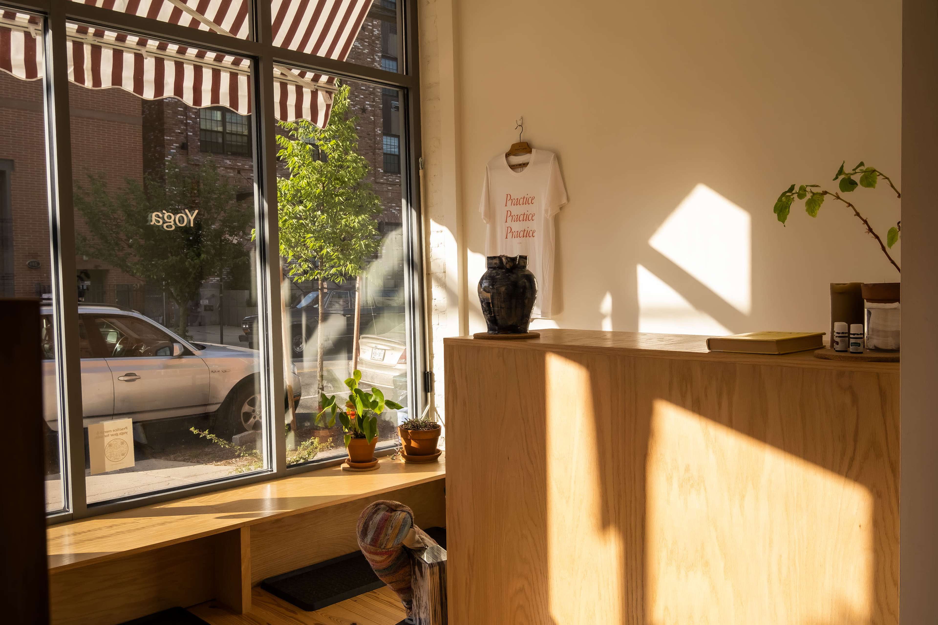 A sunlit corner of a room features a wooden counter, potted plants, and a white shirt hanging beside a window with striped awnings.