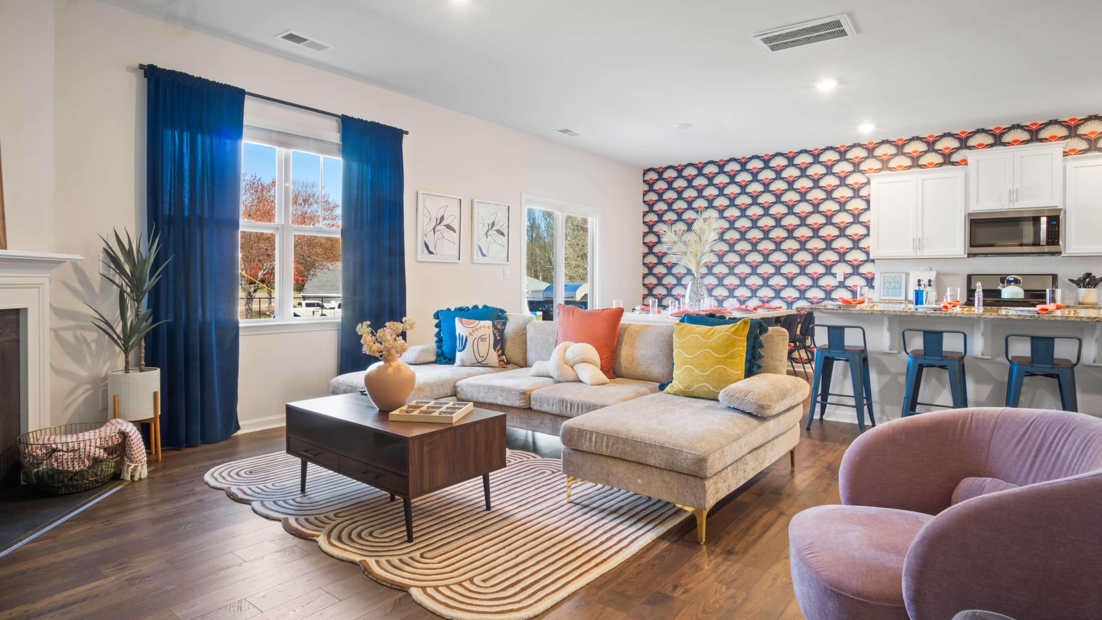 The living room features a sectional sofa, a coffee table, and kitchen bar stools, with a decorative wall and large window letting in natural light.