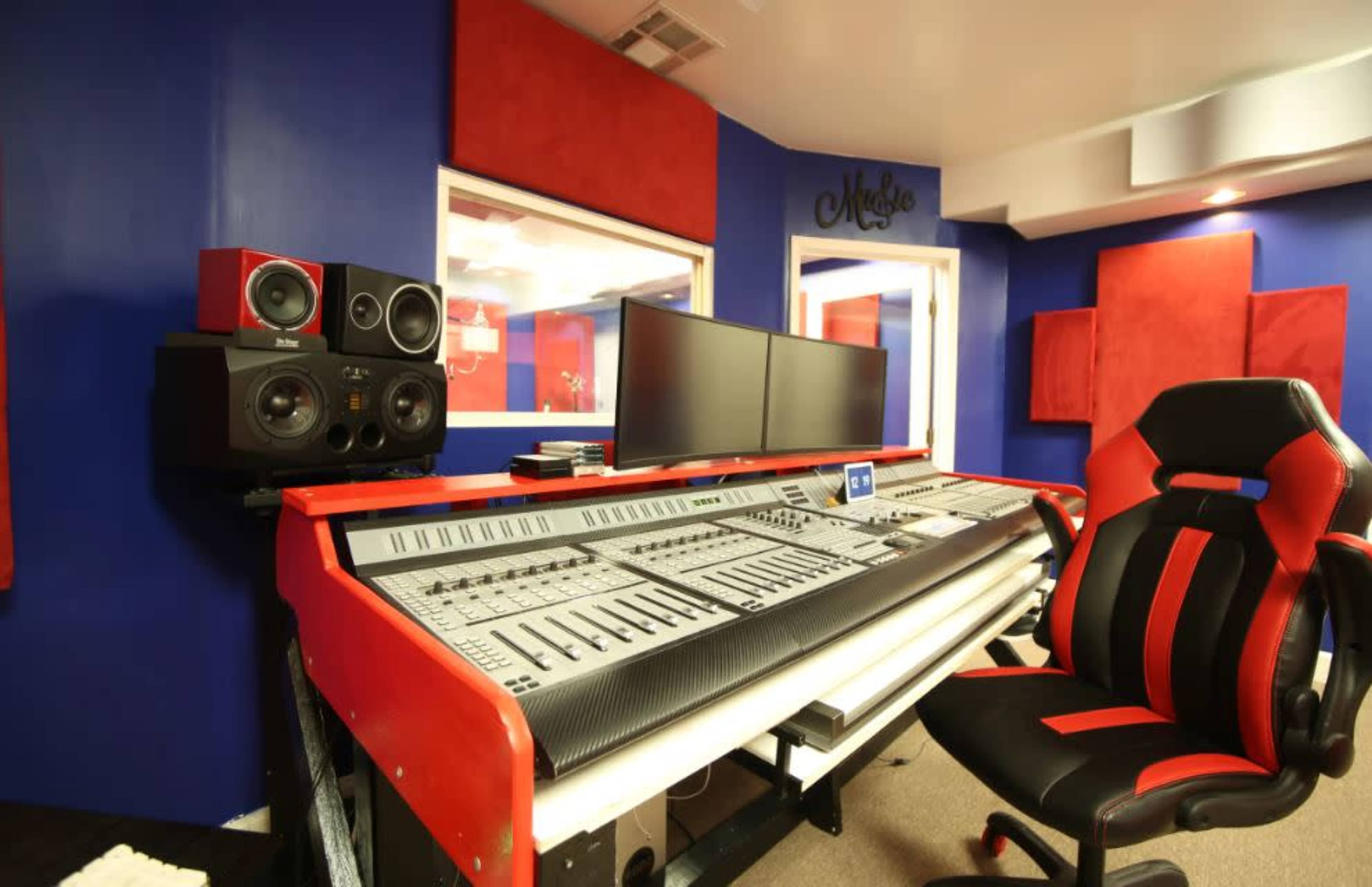The image shows a modern music recording studio with a large mixing console, two computer monitors, and acoustic panels on the walls.
