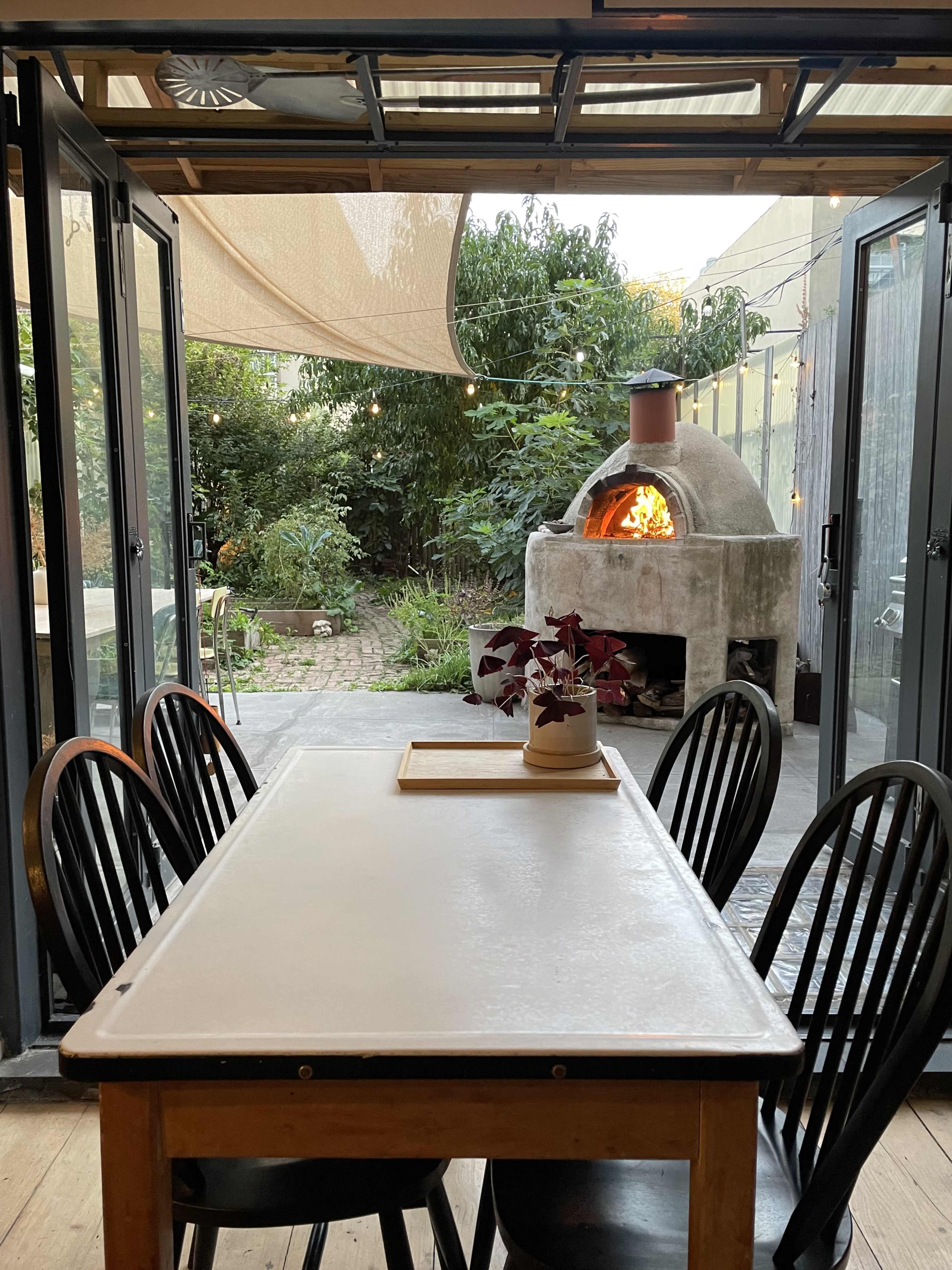 The image shows a dining table with black chairs in a room that opens to an outdoor garden featuring a stone oven with a fire burning inside.
