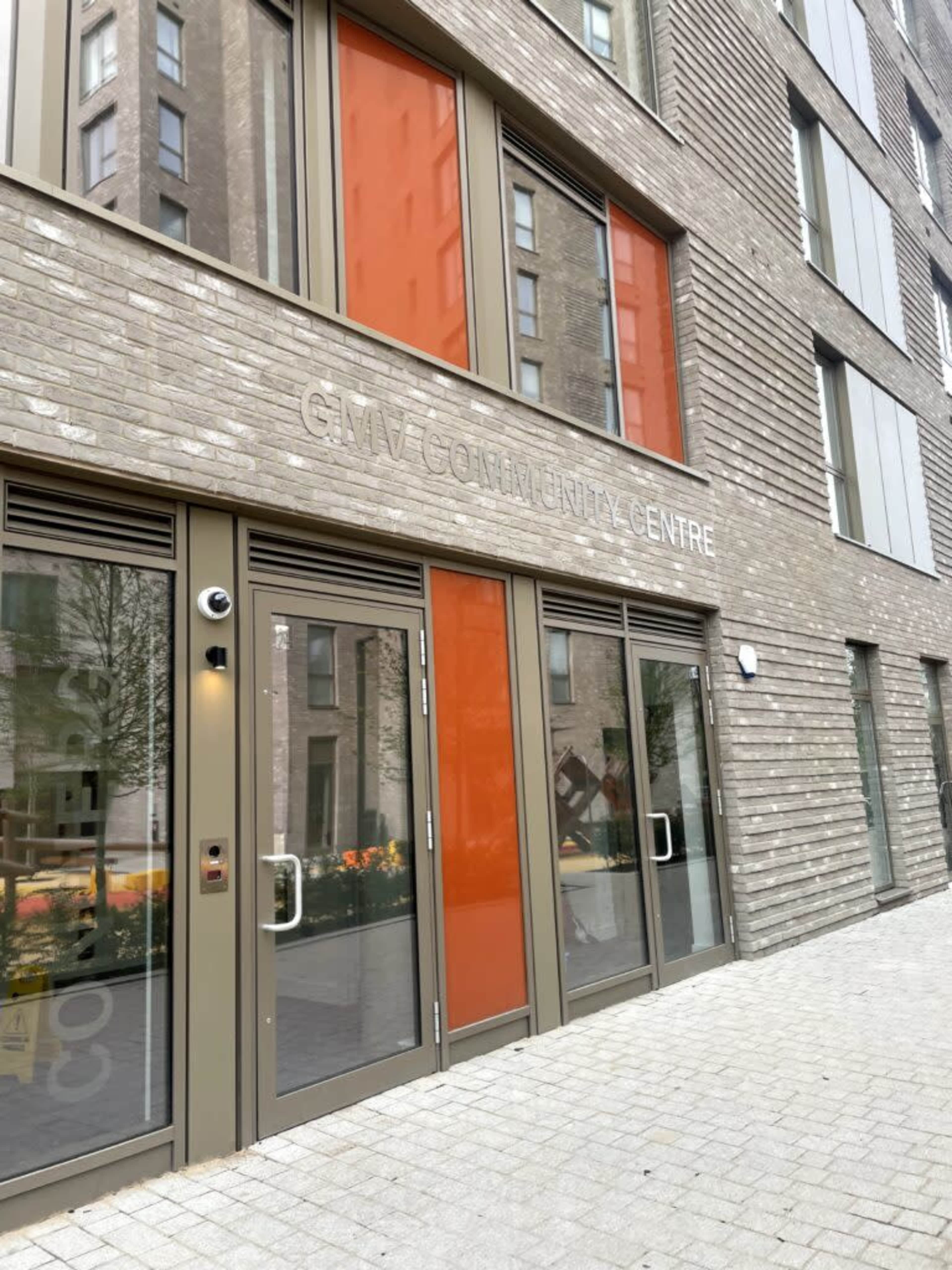 The image shows the entrance of the GMV Community Centre, featuring large glass doors and orange panels on a modern brick building.