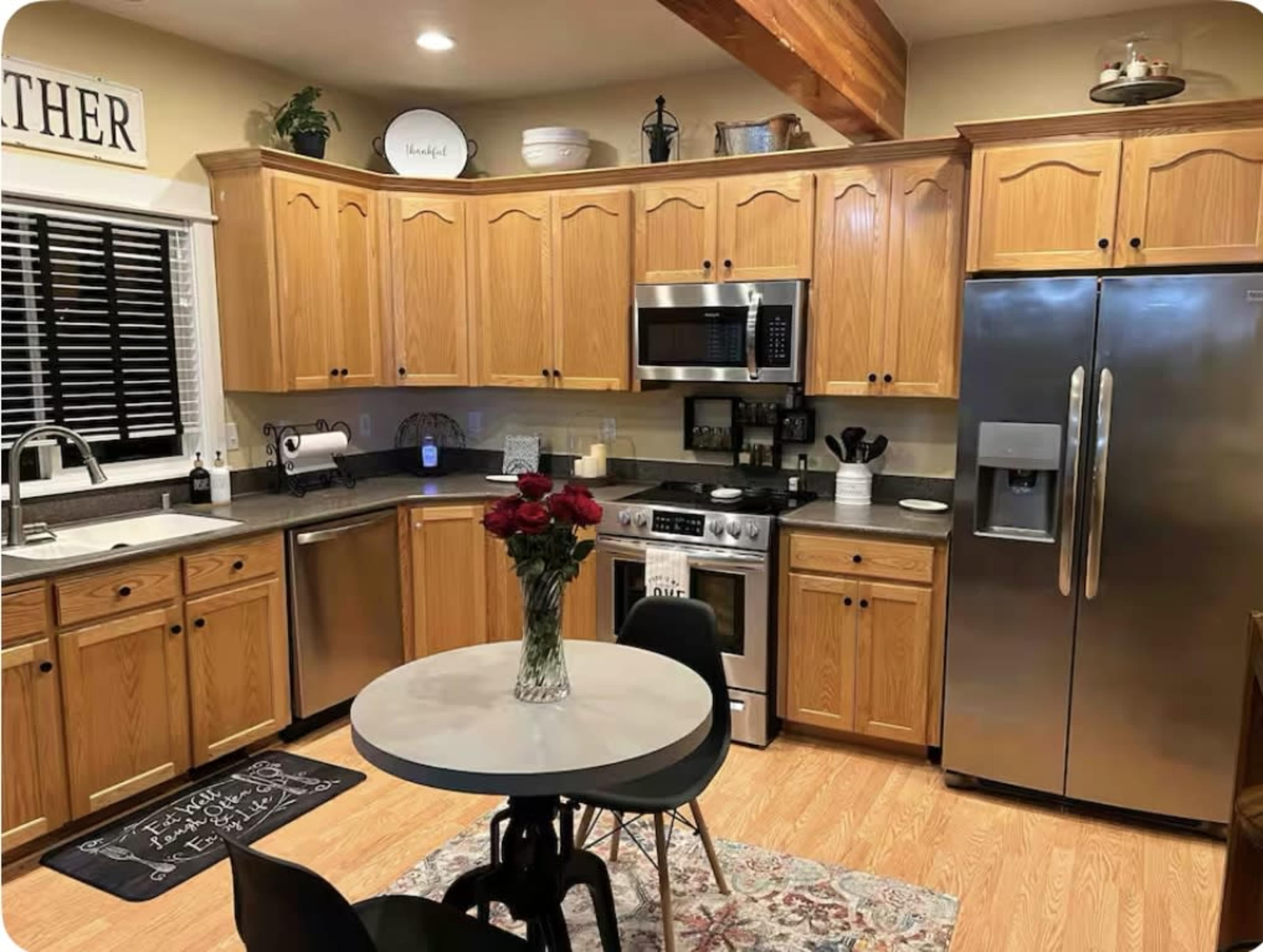 The kitchen features light wood cabinetry, stainless steel appliances, a round table with black chairs, and a bouquet of roses as a centerpiece.
