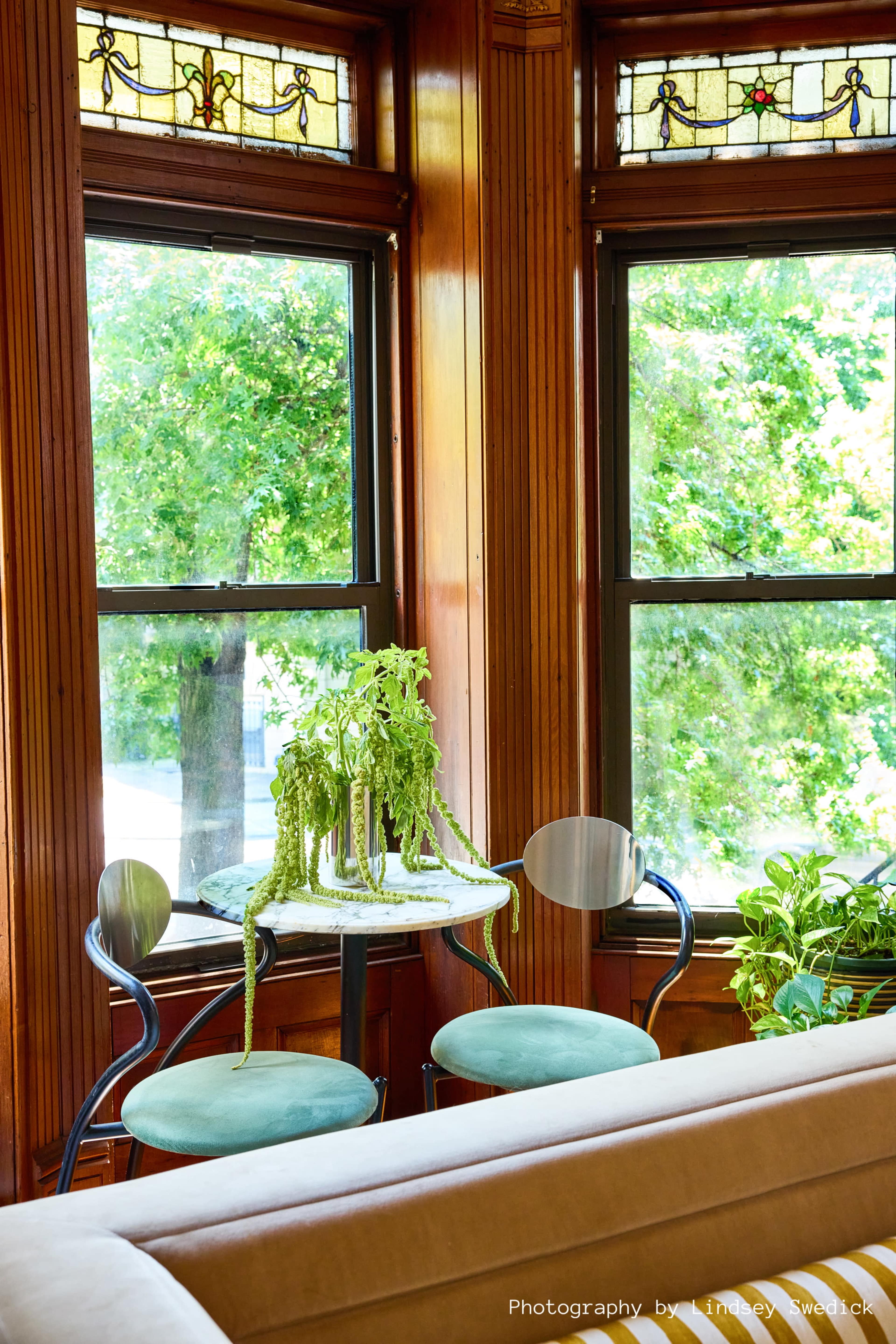 A small table with two chairs is positioned by large windows, surrounded by green plants in a wooden room.