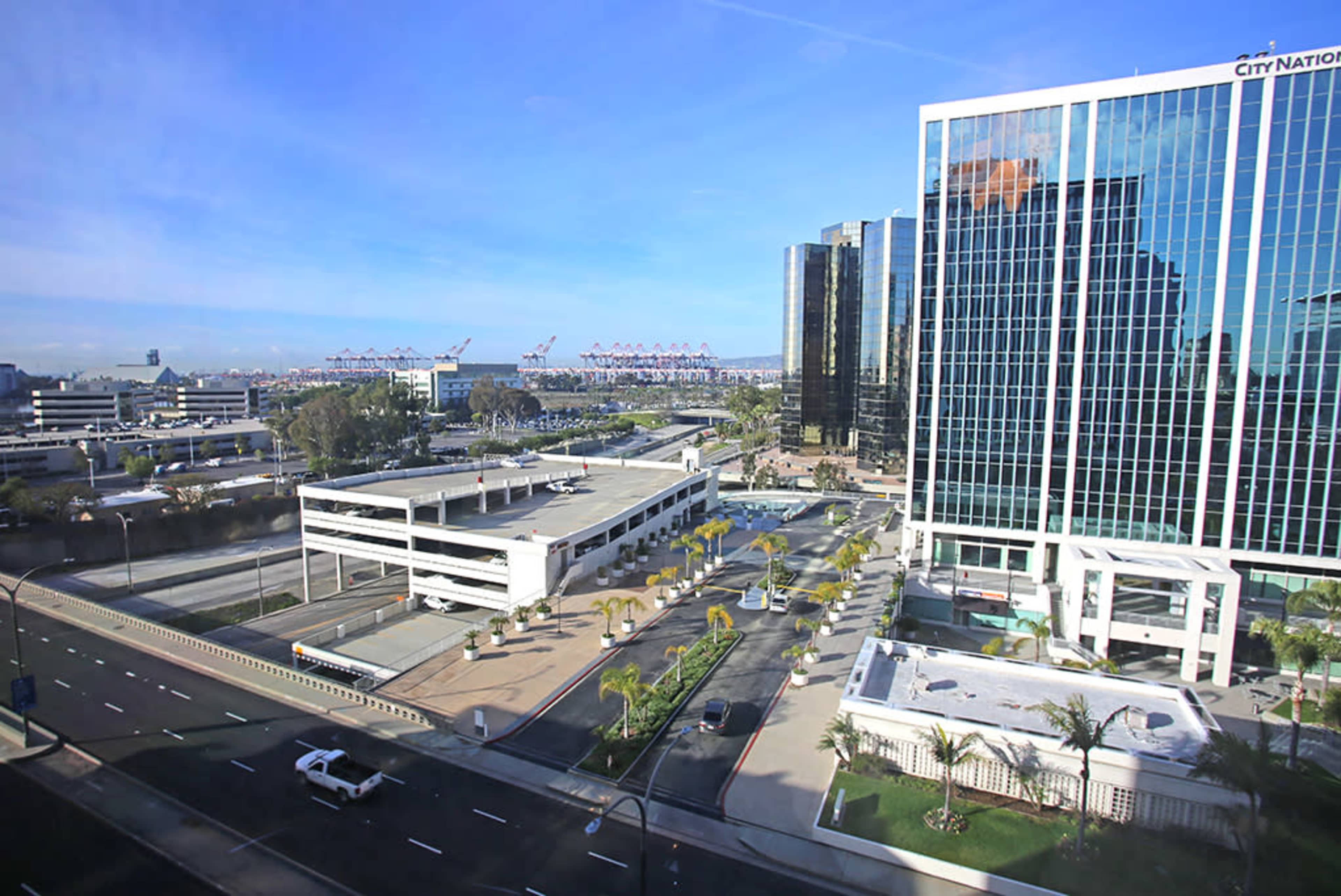 A cityscape featuring modern buildings, a parking structure, and palm trees along a road, with cranes visible in the background.