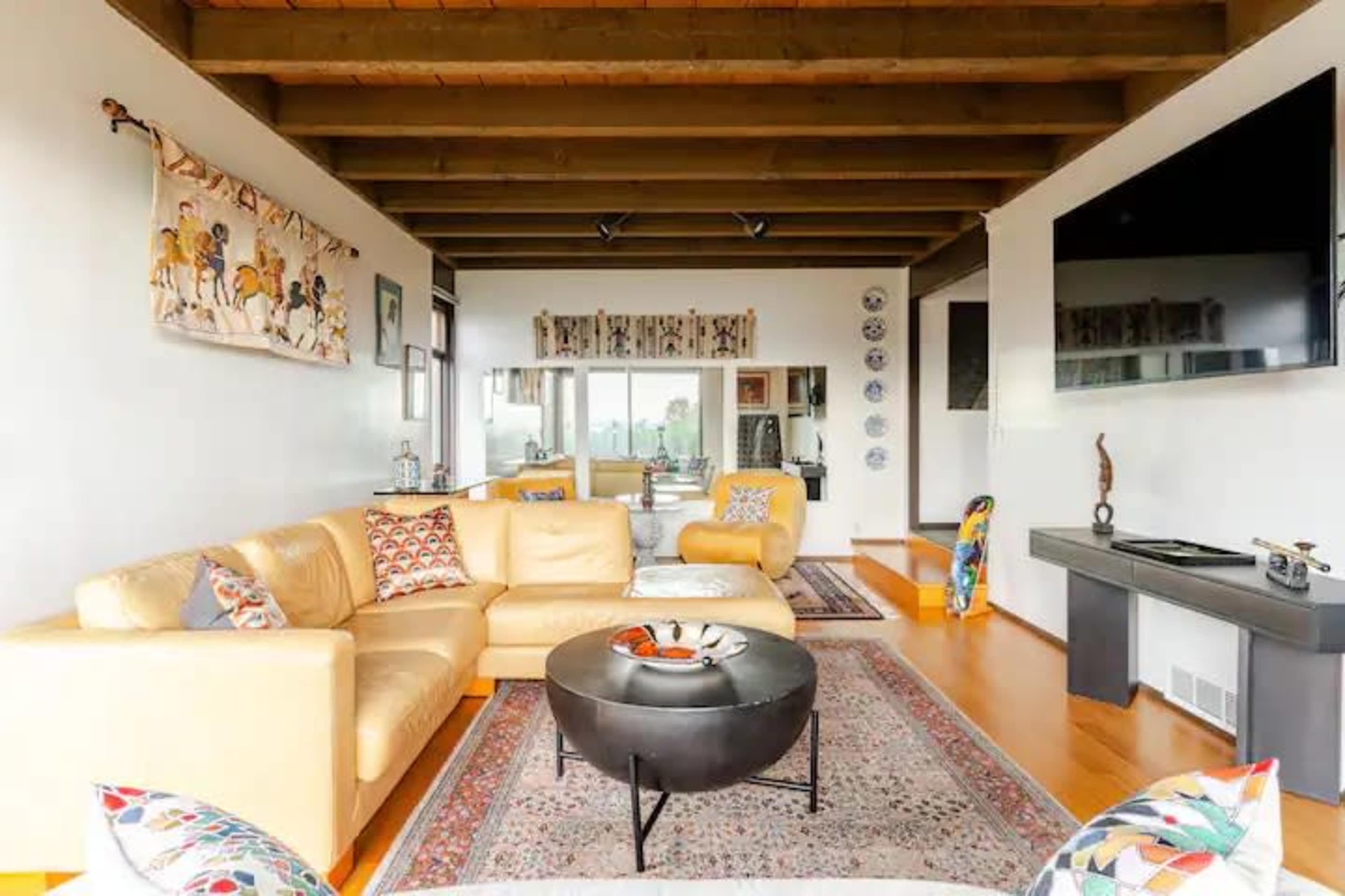 The living room features a yellow sectional couch, a round coffee table, and a large wall-mounted television, with artwork and decorative elements creating a cozy atmosphere.