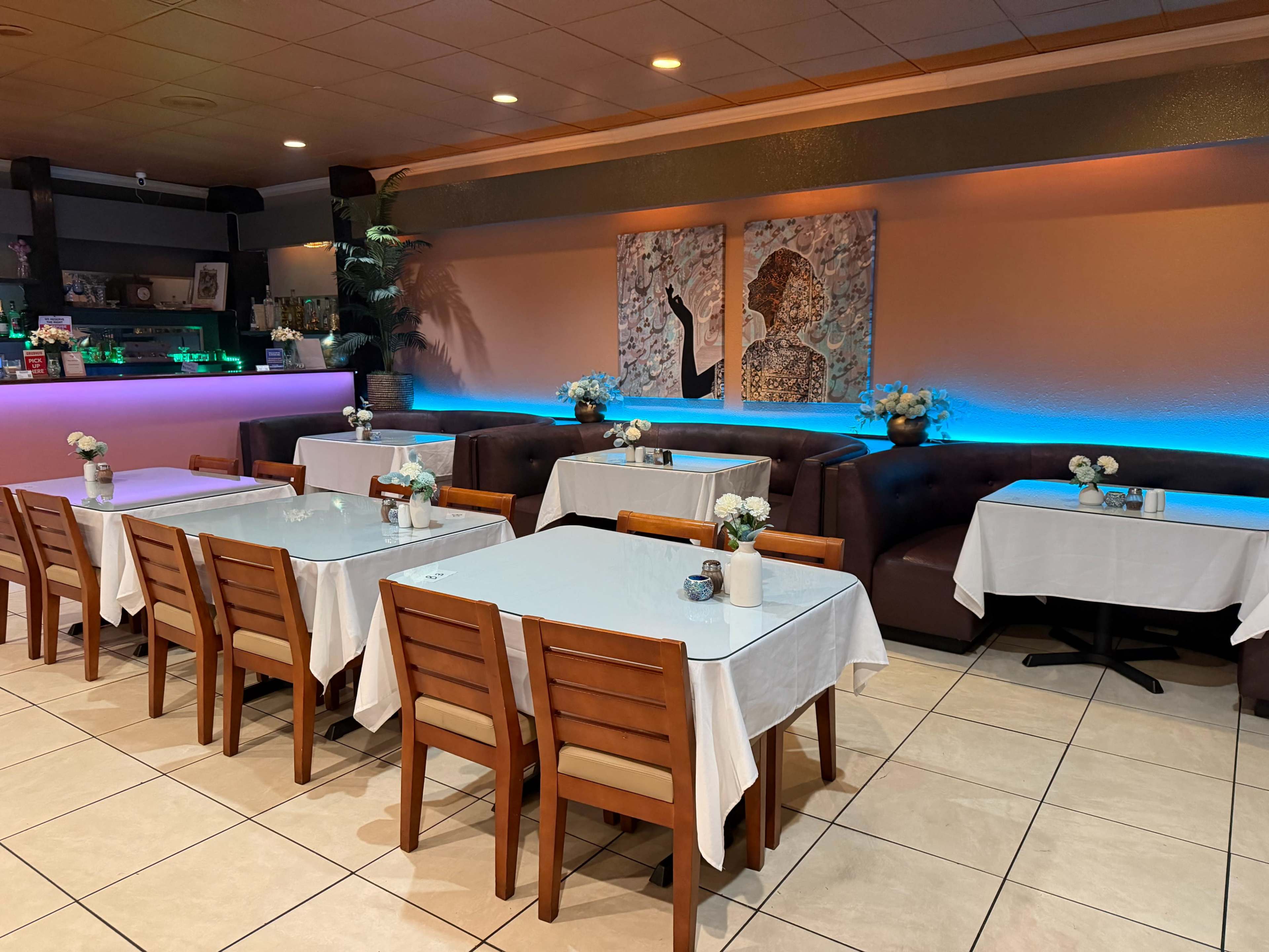 The image shows a restaurant interior with several tables set with white tablecloths and small flower arrangements, illuminated by a soft blue light in the background.