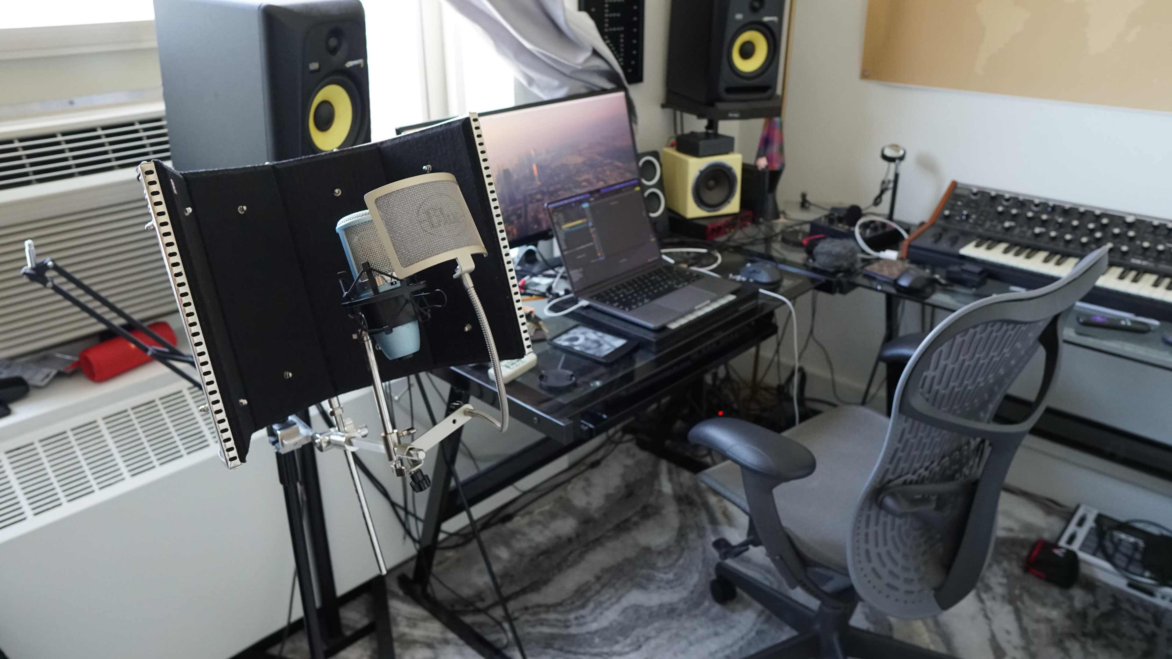 A recording studio setup features a microphone on a stand, a computer with an audio interface, and various music equipment surrounded by cables and a desk chair.